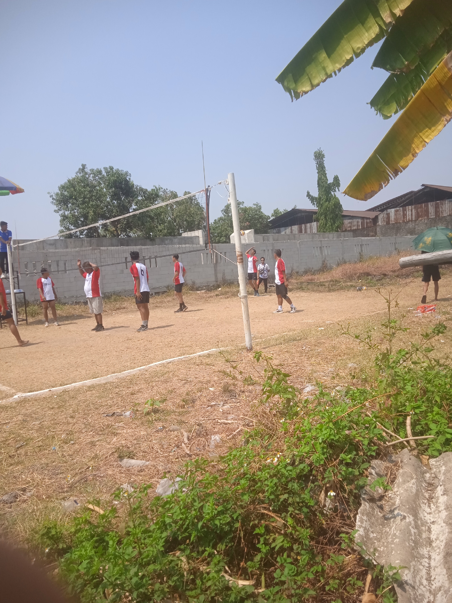 The image depicts an outdoor volleyball game in Klodran, Indonesia. Several individuals, predominantly male, are actively participating in the game on a dirt court marked with white lines. Most players are wearing red and white jerseys. The environment is dry and sunny, with sparse vegetation and trees visible in the background. A volleyball net is set up, and a volleyball is in mid-air, suggesting an active moment in the game. Buildings with corrugated roofs are present in the distance. The overall scene captures a casual sporting event in a rural or semi-urban setting.