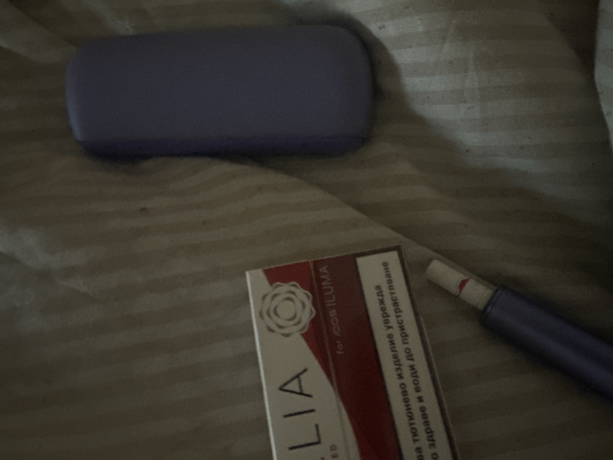 The image displays an indoor still life scene featuring several personal items on a light-colored, striped fabric, likely bedding.
The main subjects are a purple, rectangular case, a pack of tobacco sticks, and a purple tobacco heating device with a tobacco stick inserted. The case is positioned towards the top left of the frame. Below it, slightly to the right, is the tobacco stick pack, partially obscured but clearly identifiable. To the right of the pack is the purple tobacco heating device, resembling an IQOS ILUMA, with a white and red-tipped tobacco stick partially inserted. The lighting in the image is dim, suggesting either low ambient light, evening/nighttime, or an artificially lit indoor environment.
Notable details include the striped pattern of the fabric background, which has a slightly textured appearance. The objects are arranged somewhat casually. The text visible on the tobacco stick pack includes "LIA" (likely part of "TEREA"), "for IQOS ILUMA," and a health warning in Bulgarian Cyrillic script. The warning reads: "тютюнево изделие уврежда здраве и води до пристрастяване," which translates to "tobacco product harms health and leads to addiction." No specific activity is depicted, but the arrangement suggests these items are in use or readily accessible in a domestic setting. The context given is Sirakovo, Bulgaria, which aligns with the Bulgarian text on the product.