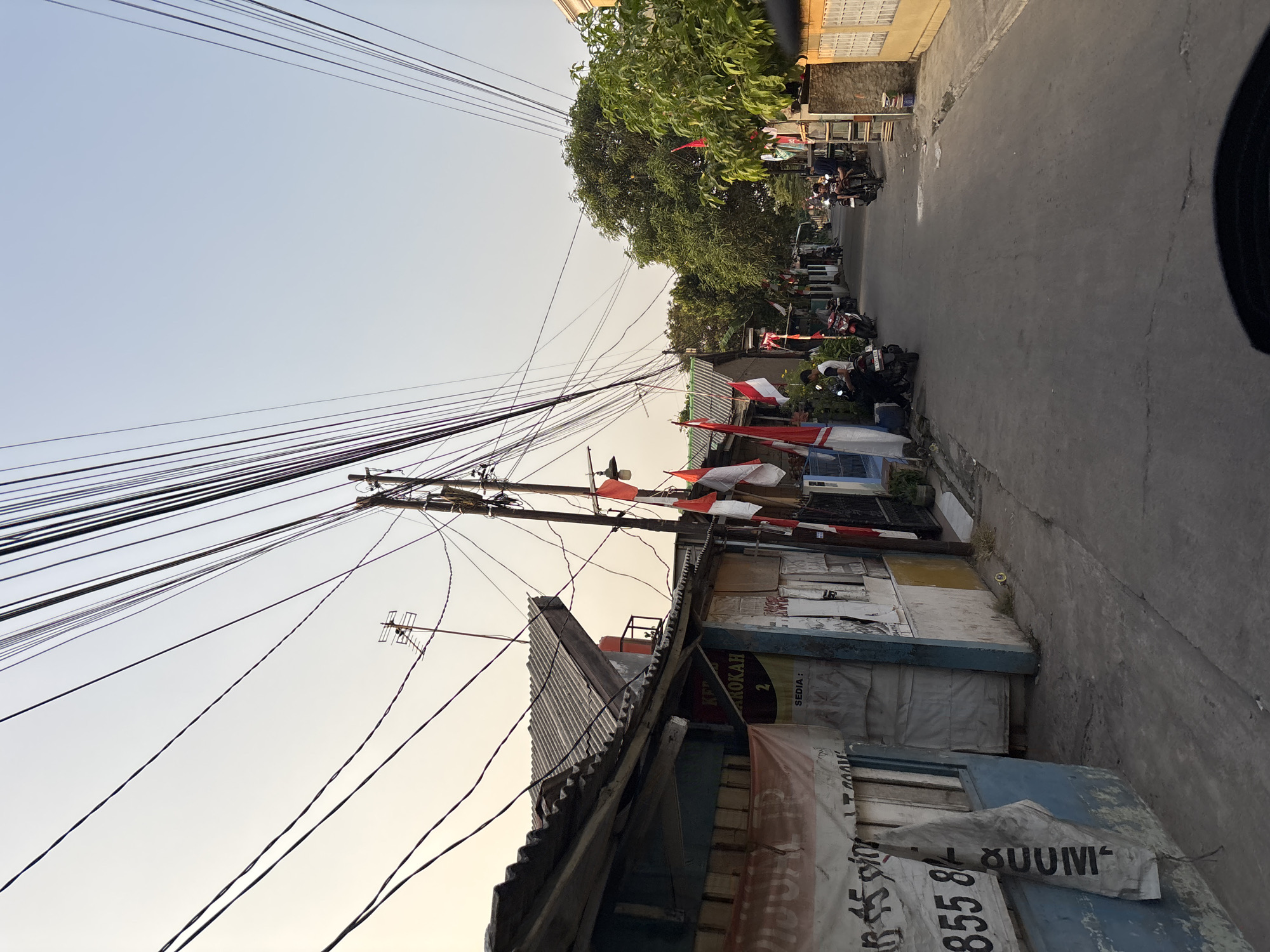 The image depicts a street scene in Bekasi, Indonesia, likely during the late afternoon or early evening, given the warm light and long shadows. The street is narrow and paved, with a row of modest, attached dwellings lining one side and trees and more houses on the other. A dense network of overhead wires, characteristic of urban infrastructure in some parts of Indonesia, dominates the upper portion of the frame, casting a web of shadows across the sky and the scene below.
Several Indonesian flags, red and white, are prominently displayed on poles and attached to buildings, suggesting a period of national celebration or remembrance. People are visible on the street, with at least two individuals on motorcycles and others standing near their homes. The buildings themselves appear to be constructed from a variety of materials, including corrugated metal roofing, painted walls, and what looks like salvaged materials. Banners and signs are affixed to some of the structures, some of which are partially obscured or weathered.
The general impression is one of a lived-in, functional urban environment, perhaps a neighborhood with a strong sense of community, evidenced by the flags and the communal nature of the street. The scene is bustling but not overly crowded, with everyday activities taking place. The presence of multiple motorcycles indicates a common mode of transportation in this locale. The sky is clear and light, with a gradient from pale yellow near the horizon to a soft blue above, hinting at either sunrise or sunset.