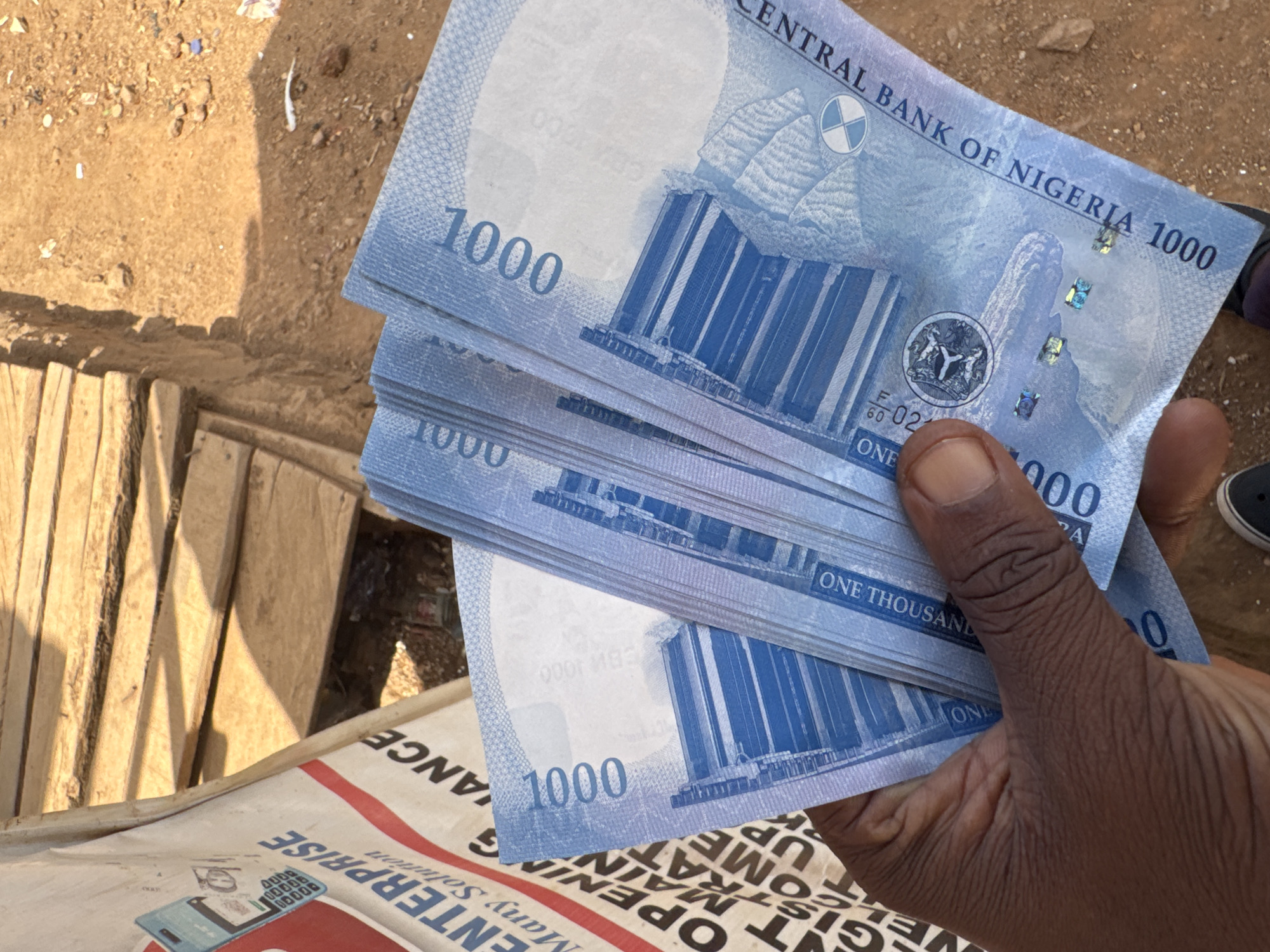 A close-up, high-angle outdoor shot features a dark-skinned hand holding a fanned stack of new Nigerian 1000 Naira banknotes. The banknotes are predominantly light blue and appear crisp. The front of one note clearly displays "CENTRAL BANK OF NIGERIA 1000" at the top, the denomination "1000" on the left, and an image of a building. Security features are visible on the note. The reverse of another note shows the words "ONE THOUSAND" printed horizontally, and mirrored text like "CBM 1000" is also faintly discernible.
The background primarily consists of rough, sun-drenched sandy ground with scattered small debris. To the bottom left, several wooden planks are visible, likely part of a makeshift structure or walkway. Below the hand and planks, a white bag or sack is partially visible, featuring red and black text. Visible text on the bag includes "ENTERPRISE", "Many Solution", "OPENING", "MAIN", "GIOME", "UP", and "000L ANCE" (some fragmented or upside down).
The scene is brightly lit by natural daylight, casting clear shadows on the ground and indicating daytime. The setting appears to be an outdoor, informal environment, possibly a market or street in Kurunduma, Nigeria. The image captures the act of holding or presenting a significant amount of local currency.