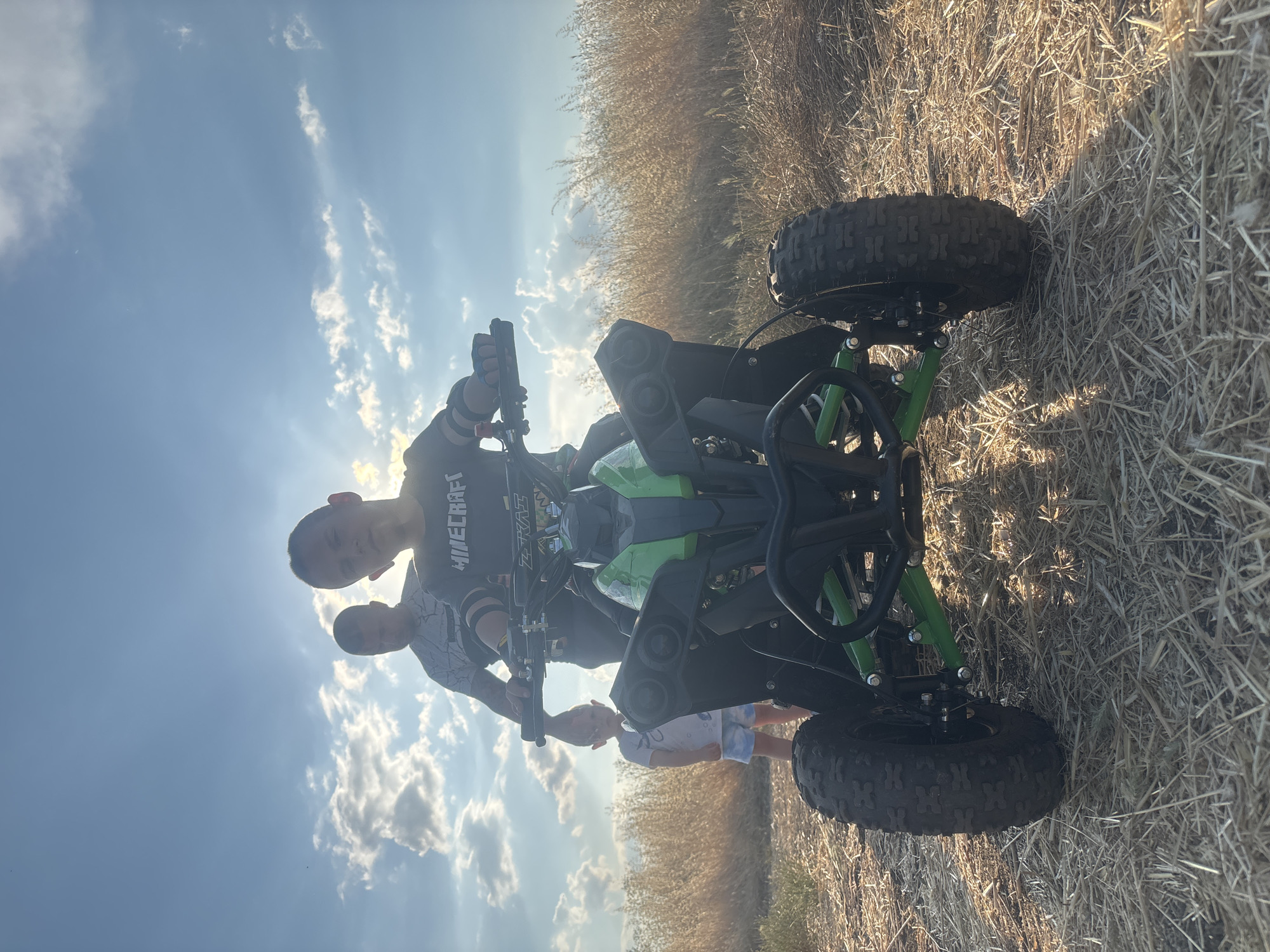 In a dry, grassy field, bathed in the golden light of a setting sun with a partly cloudy sky, two boys and an adult stand near a green and black ATV. The ATV is positioned in the foreground, its large, knobby tires resting on the dry earth. A young boy, wearing a black long-sleeved shirt with "MINECRAFT" printed on it, sits on the ATV, his hands on the handlebars, looking towards the camera. Behind him, an adult, wearing a grey t-shirt with a white patterned design, stands with one hand on the ATV. To the left of the ATV, a younger child in a white t-shirt and blue shorts stands, looking towards the adult. The background consists of dry, tall grass and a bright sky with visible sun rays breaking through clouds. The scene does not provide any specific indicators of a location within the city of Burgas, Bulgaria.
