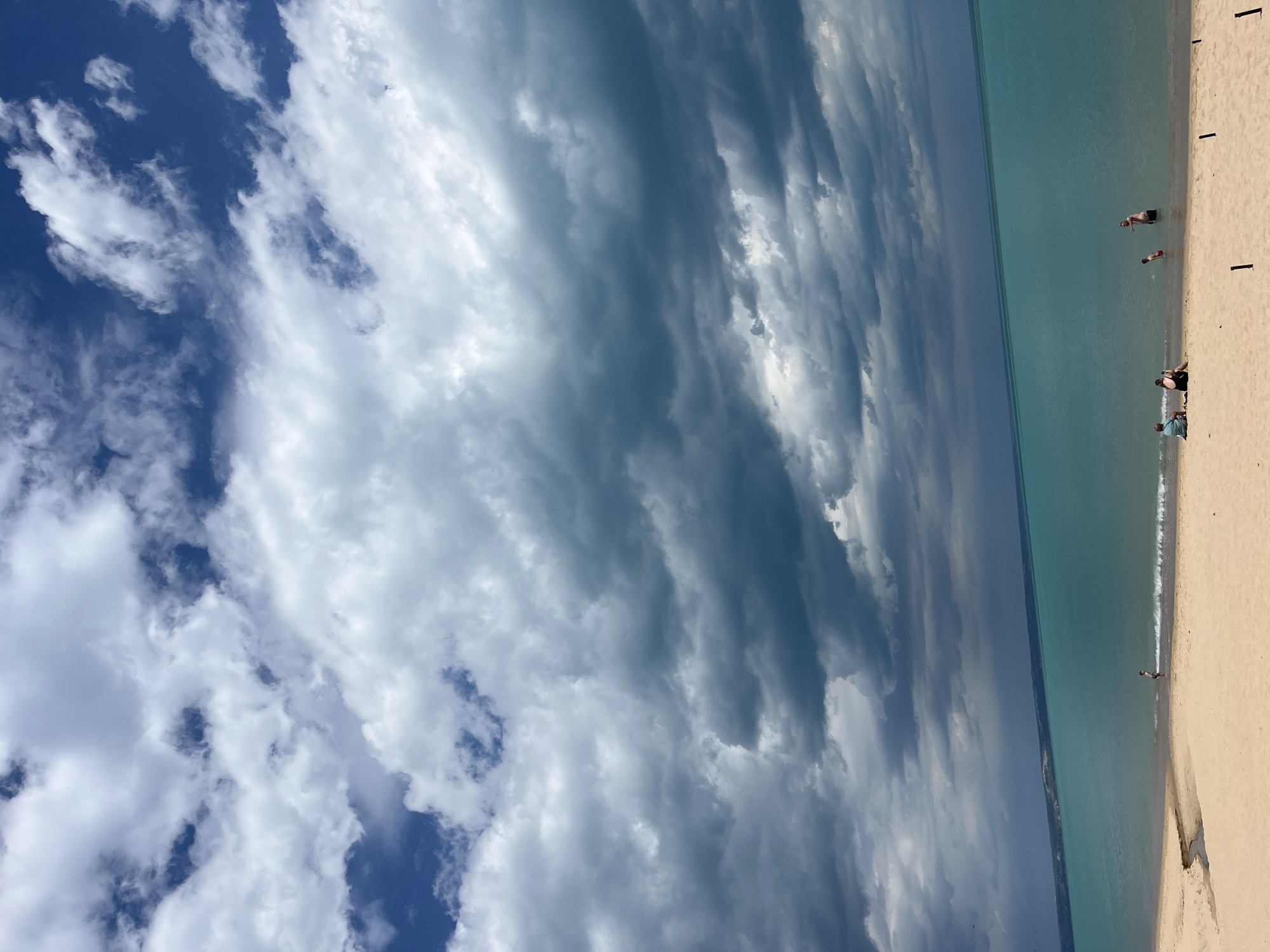 The image shows a beach scene in Varna, Bulgaria. The view is oriented vertically, with the sky dominating the left side of the frame. The sky transitions from patches of blue with white, puffy clouds to a dense layer of dark, striated clouds covering the center of the frame. The sea is to the right, a clear turquoise color. A sandy beach lines the right edge of the frame. Several small groups of people are on the beach or in the water, including a group sitting on the sand near the bottom of the frame, and two individuals standing in the water further up the beach. T-shaped structures are standing on the beach.