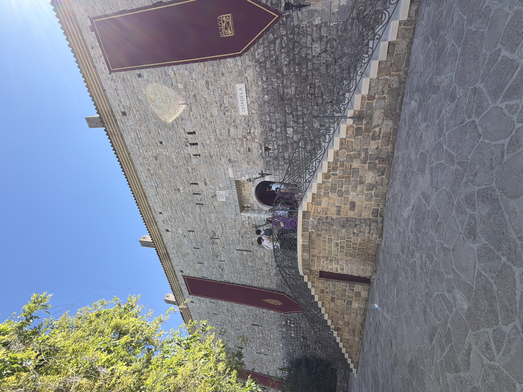 The image shows a section of the Citadel in Budva, Montenegro. A large stone building with a burgundy banner that reads "Budva" in Cyrillic is visible. A stone staircase with a wrought iron railing leads up to an arched doorway where several people are standing. The building's wall is made of light-colored stones. The ground is paved with gray stone slabs. Lush green foliage is in the lower-left corner, and a clear blue sky is above.