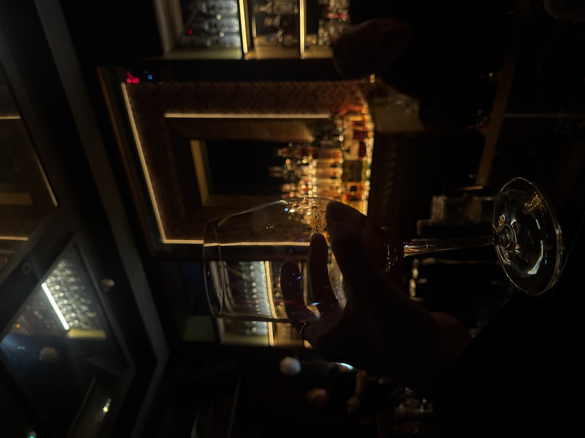 The image captures a dimly lit bar in Dubai, United Arab Emirates. A hand holds a wine glass in the foreground, obscuring a portion of the background. The bar area is defined by rows of illuminated shelves stocked with various bottles, likely alcoholic beverages. The lighting creates a tunnel-like effect, with a sense of depth extending into the background.