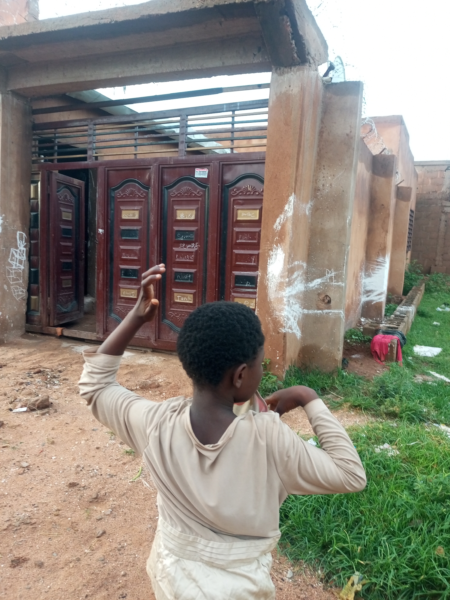 In the city of Tempure, Nigeria, a young Black individual stands with their back to the camera, facing a large, ornate, maroon-colored gate. The gate has multiple panels with gold detailing and appears to be partially open. The individual is wearing a light-colored, long-sleeved shirt and a lighter-colored skirt or pants with a pleated waistband. Their right arm is raised, with their hand open and fingers spread. The ground in front of the gate is dirt and sand. To the right of the gate, there is a building with concrete pillars that have white markings on them, resembling chalk drawings. Lush green grass grows to the right and behind the building, with some debris scattered in the grass. The overall environment suggests an outdoor setting in a residential or semi-developed area.