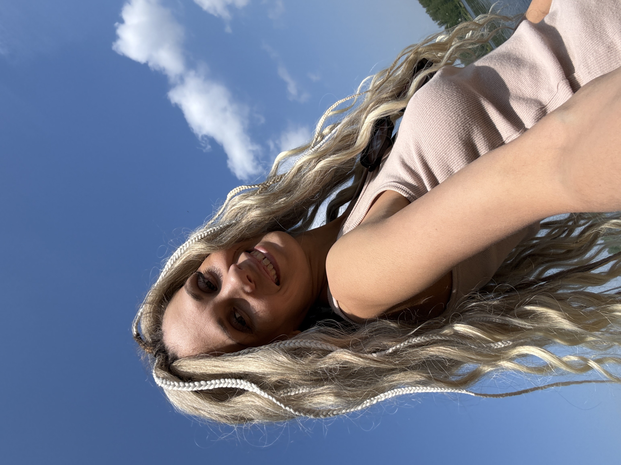 A low-angle, rotated image shows a woman with long, wavy blonde hair interspersed with white braids smiling towards the camera against a backdrop of a bright blue sky with scattered white clouds. She is wearing a light pink, textured top. The photograph appears to be taken outdoors in the city of Rusanovo, Russia near a body of water, with a glimpse of green trees visible in the upper right corner.