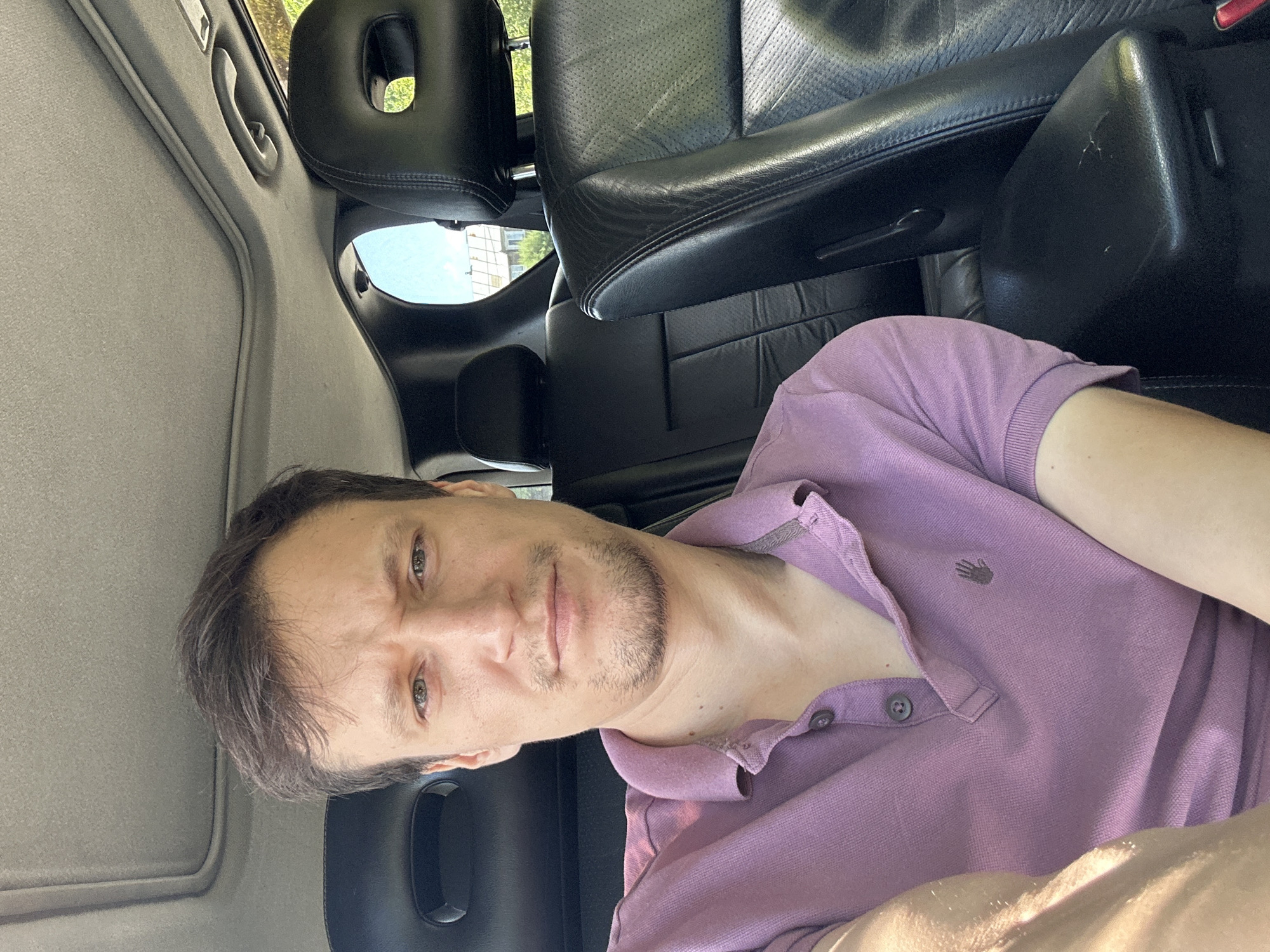 The image shows a man sitting in the driver's seat of a car, with the camera positioned at a slightly upward angle. He has short, dark hair and is wearing a purple polo shirt. His expression is neutral, with his eyebrows slightly furrowed. The interior of the car is visible, including black leather seats and a sunroof. Outside the car, a bright, sunny day is suggested by the light. The location of Makiv, Ukraine, is not discernible from the visual information in the image.