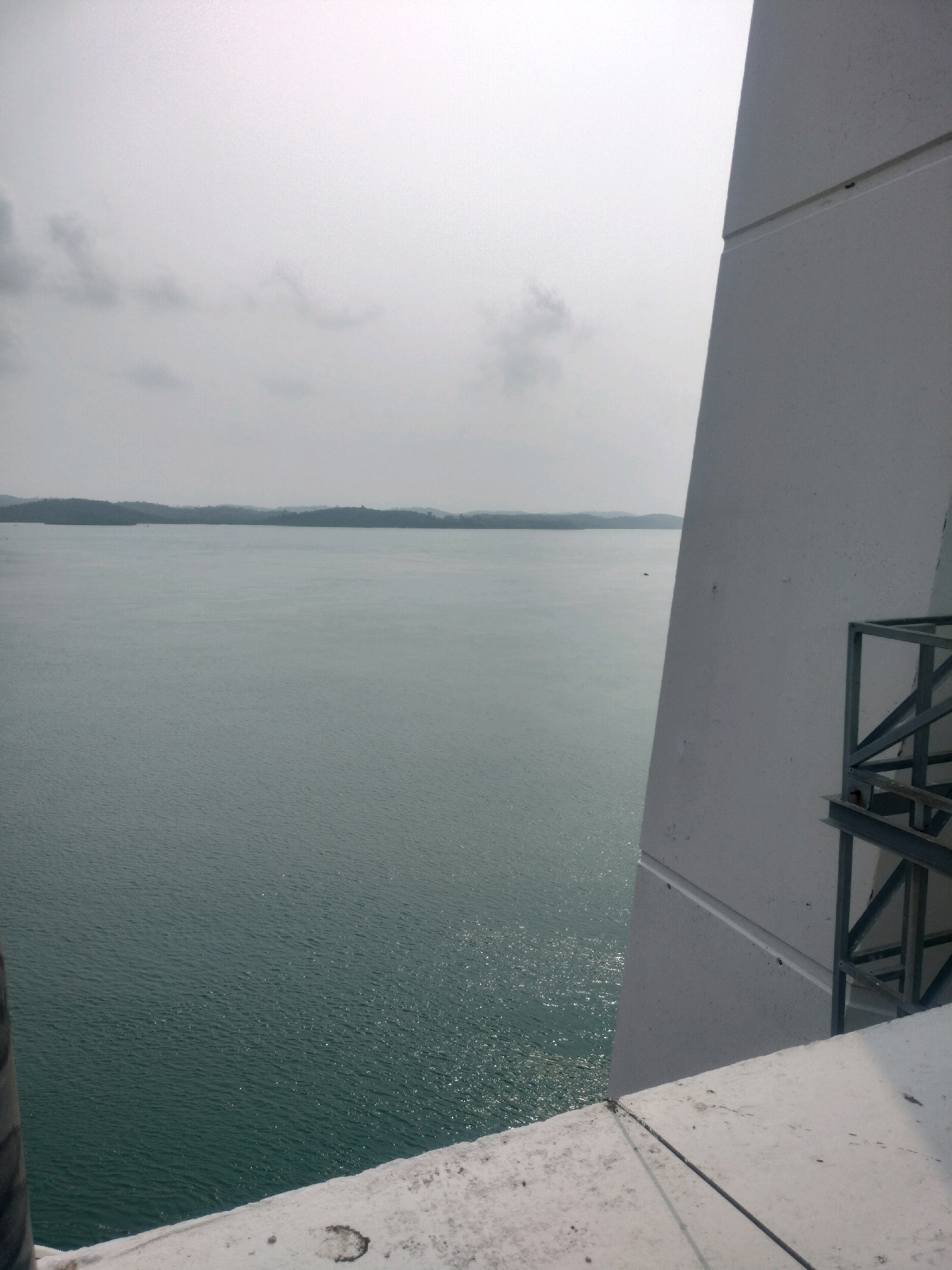 The image shows a view of the sea from a high vantage point, likely a bridge or a tall structure, in Batam, Indonesia. In the foreground, a large, white, angled structure dominates the right side of the frame. This structure appears to be made of concrete and has visible panel lines. To the right of this structure, a metallic railing or walkway is attached. The sea stretches out to the horizon, meeting a hazy sky with scattered clouds. The water is a deep green-blue and appears calm with small ripples reflecting sunlight. In the distance, a line of green, hilly islands forms the horizon. A tiny dark speck, possibly a boat, is visible on the water. The bottom of the frame shows a white, textured surface, likely part of the structure the photo is taken from, with some dirt or debris. The overall impression is one of a vast, open seascape under an overcast sky.