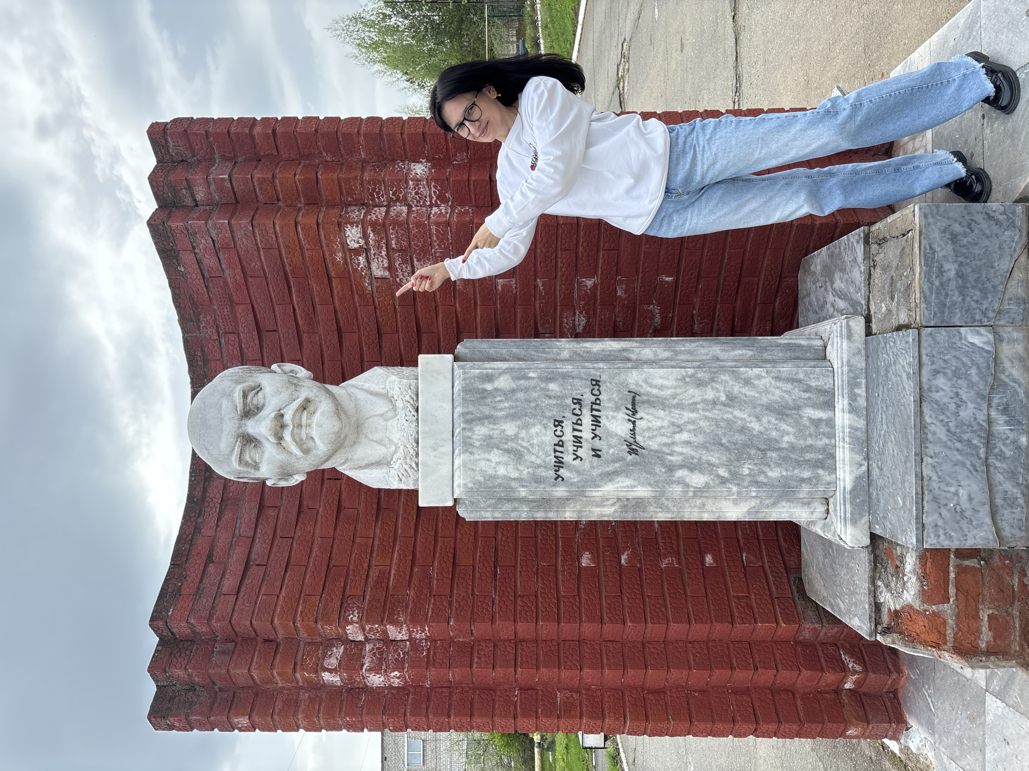 A woman in a white sweatshirt, blue jeans, and black shoes is standing on a gray concrete or marble base, pointing her right index finger at a bust of Vladimir Lenin. The bust is white marble, and sits on a tall gray marble or concrete pedestal in front of a large, stepped red brick wall. The inscription on the pedestal is in Russian. The location is outside, likely in a public area or park, in the city of Chaikovsky, Russia. The background shows a cloudy sky, a grassy area, and a building. The photo has been rotated.