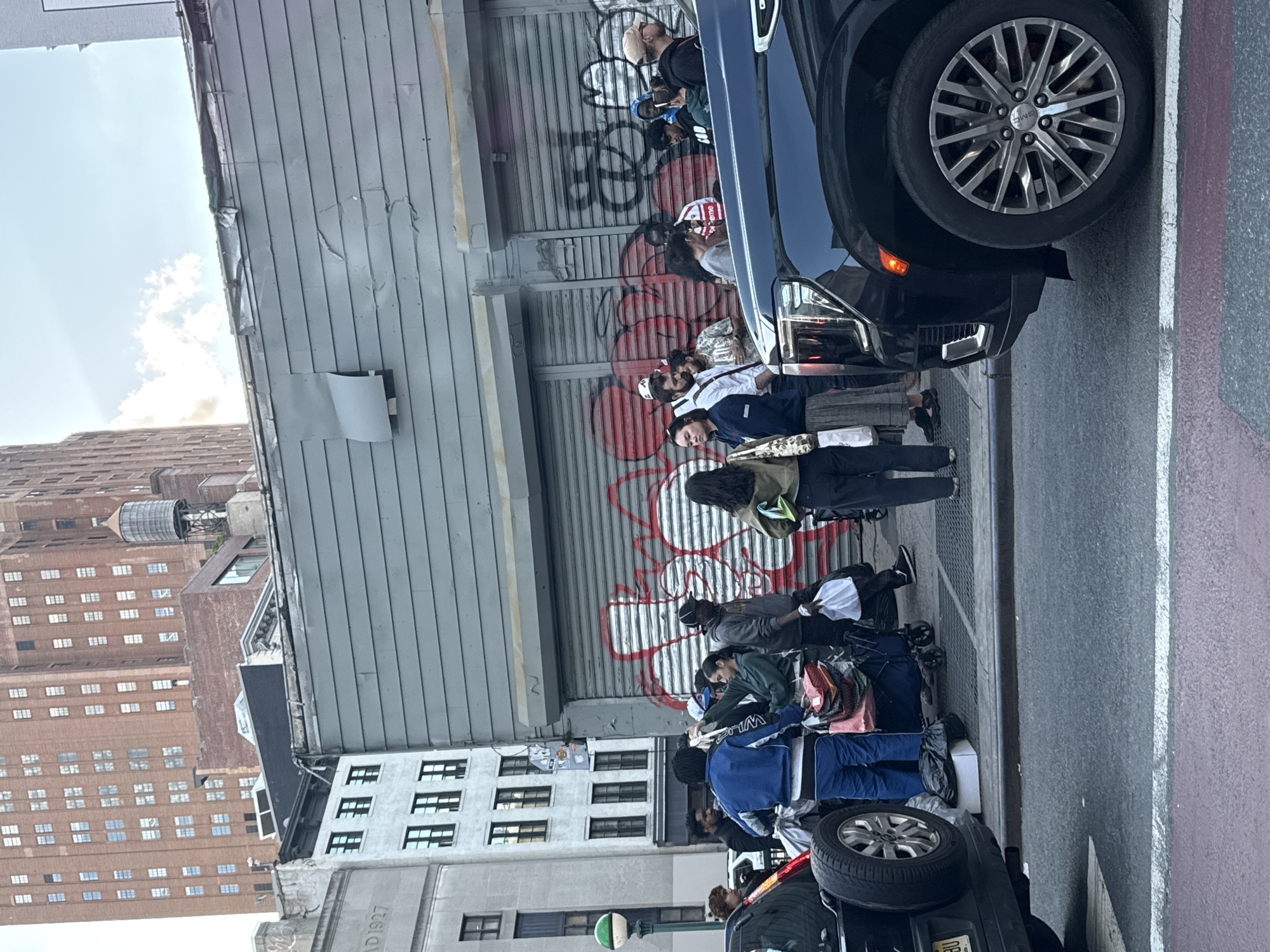 The image shows a group of people standing in line on a sidewalk in New York City. The line is positioned in front of a closed metal shutter door covered in graffiti. A dark-colored SUV is parked partially on the sidewalk at the head of the line. Adjacent to the queue and across the road are older buildings, one of which has "AD 1927" inscribed on its facade. In the background, a tall brick building with a water tower on its roof is visible. The overall scene appears to be an urban environment.
