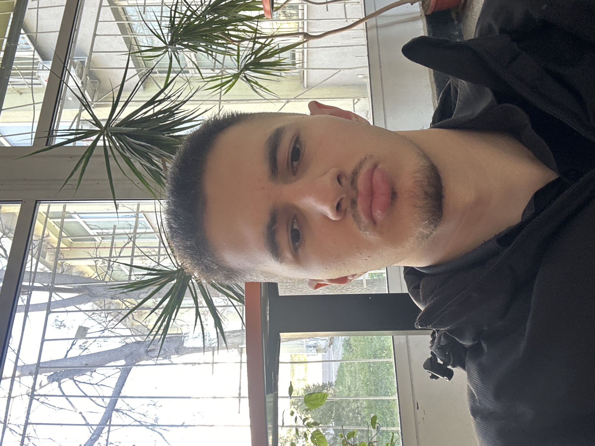 A close-up selfie of a young man with short dark hair and a light complexion. He is wearing a black jacket. The man is facing the camera with a slightly pursed lip expression.
Behind him, there is a window with a metal grid. Outside the window, there is a building and greenery visible. A potted plant with long, thin leaves is positioned near the window. Based on the architectural style and the vegetation, it is likely in the city of Sofia, Bulgaria.