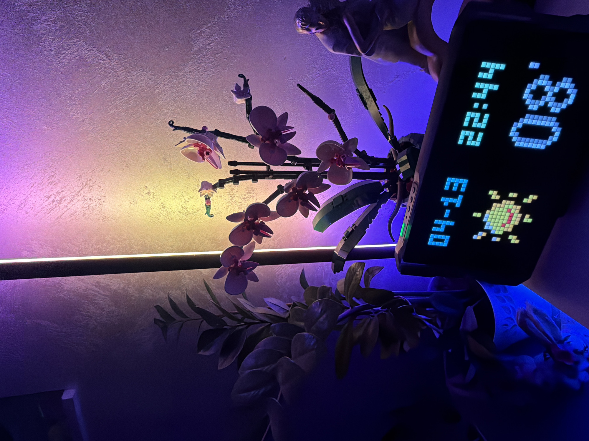 The image depicts a close-up shot indoors, under dim, artificial lighting with hues of purple, blue, and yellow. The primary subjects are: a Lego orchid (partially in focus), a digital clock with an 8-bit pixel art display showing "14:22" and "80" with a space invader graphic, a figurine partially visible at the top, and a potted plant with dark green leaves. The background shows a textured wall illuminated by a linear light fixture. It is impossible to determine the location is Varna, Bulgaria without further information.