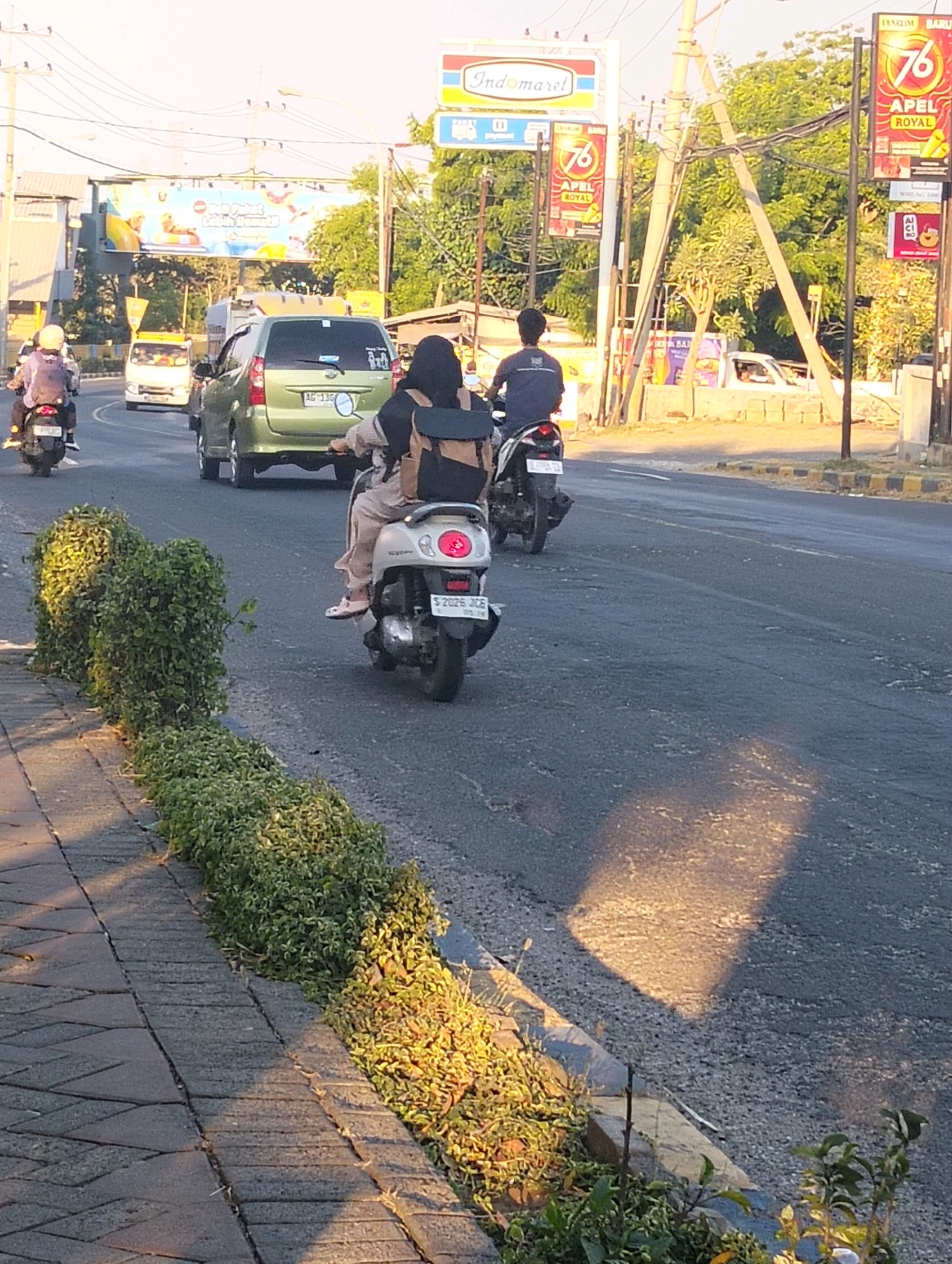 The image captures a street scene in Tunggul, Indonesia, likely during late afternoon given the long shadows. The foreground features a textured, paved sidewalk bordered by a low hedge of green and yellowing plants, which extends along the edge of the asphalt road. The road itself is a multi-lane thoroughfare with some visible cracks and patches, and sunlight casts a rectangular shadow on its surface.
Several vehicles are present. Closest to the camera, a woman wearing a headscarf and light-colored clothing is riding a scooter. She has a backpack on. Behind her, another person is riding a scooter, also with a backpack. Further down the road, a green minivan is moving away from the camera, with a license plate that appears to read "AG 130". To the left of the minivan, another scooter is visible from behind, with a person wearing a helmet and a backpack. In the distance, a white truck is also on the road.
Overhead, electrical wires and poles are prominent. Several billboards are erected along the roadside. One large billboard displays colorful imagery, possibly advertising a tourist destination. Other smaller billboards show advertisements for "Indomaret" and "APEL ROYAL" with the number "76". Lush green trees line the background, suggesting a tropical or subtropical environment. The overall impression is of a busy urban street with ongoing traffic.
