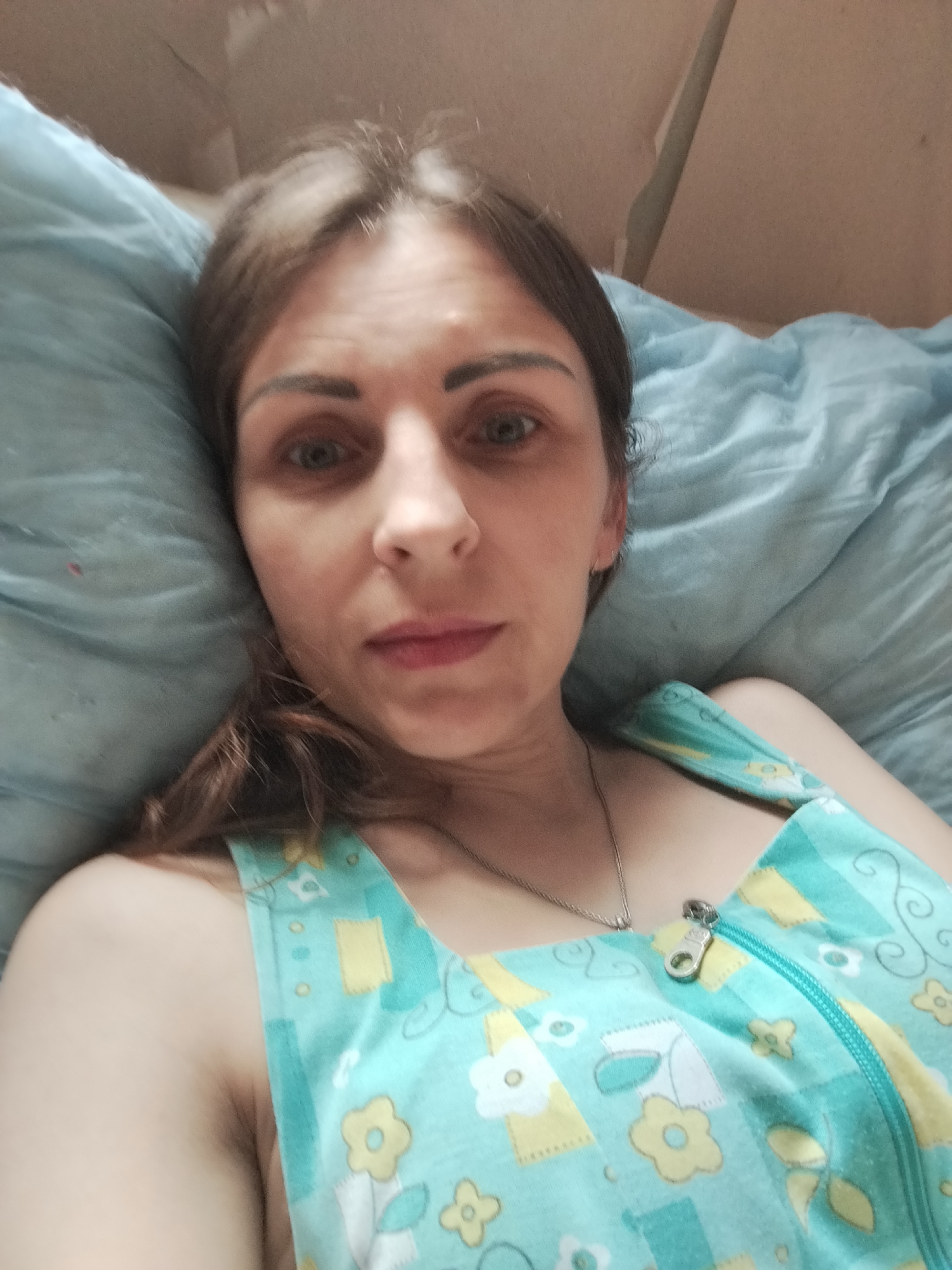 A woman is lying on a bed, looking at the camera. She has brown hair and is wearing a light blue and yellow floral patterned top with a zipper. The background is a light blue blanket and a light brown wall.