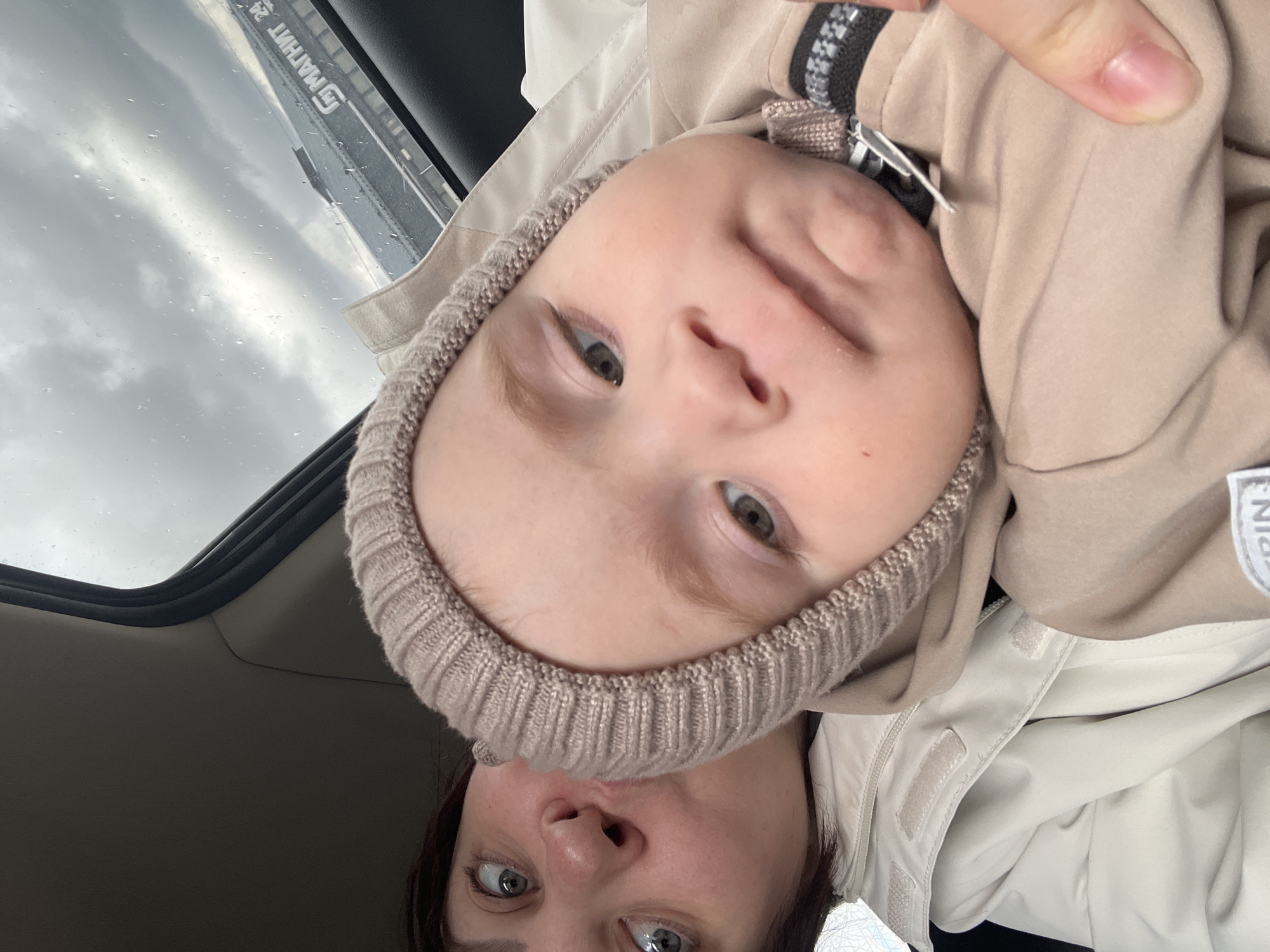 This rotated close-up portrait features an adult and a child, likely taken as a selfie inside a car. The child, positioned in the upper part of the frame (when oriented correctly), wears a light brown knitted hat covering their ears and a beige jacket. They have prominent dark eyes and a neutral, curious expression, looking directly at the camera. Below the child, only the eyes, nose, and forehead of an adult with dark hair are visible. The adult's eyes are wide and focused on the camera, and they are wearing a light beige or off-white jacket with visible velcro closures on the front placket.
The setting appears to be the interior of a vehicle, with a dark car roof lining visible. Through a car window, an overcast sky with visible raindrops suggests cloudy or rainy weather. A reflection or view of a building is visible on the window glass, featuring white Cyrillic text that reads "МАГНИТ 24". This text strongly indicates a Russian location, aligning with the provided context of Protasy, Russia, as Magnit is a major Russian supermarket chain. The image captures an intimate moment, likely during daytime, showing the two individuals observing the camera from within the car.