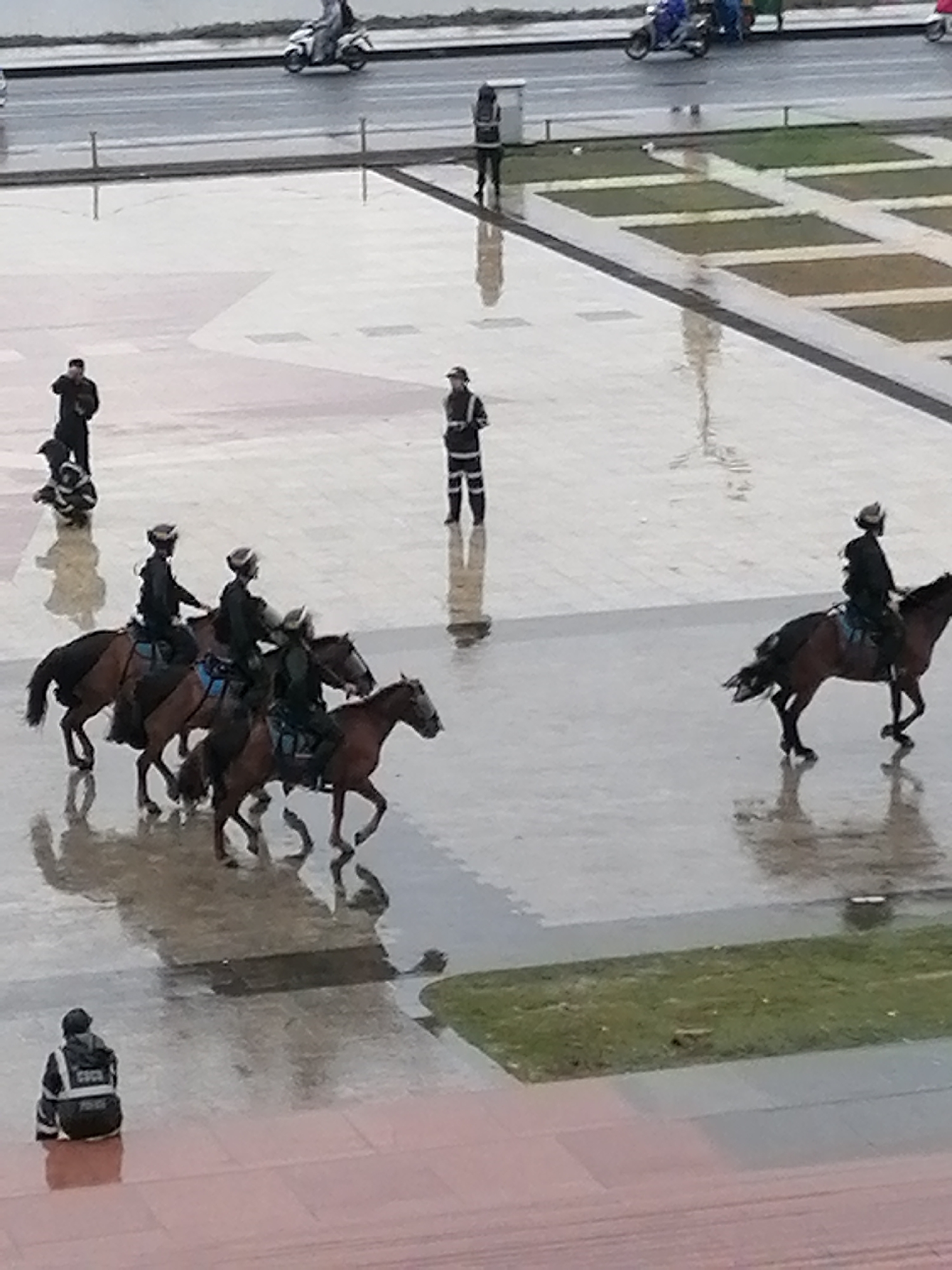 The image depicts a scene in Da Lat, Vietnam, on a wet day. In the background, several people are seen near motorcycles and scooters on a paved road. In the middle ground, four individuals on horseback, dressed in dark uniforms and helmets, are riding across a wet, reflective paved area. One person in a similar uniform is standing alone in the background, also in the paved area. In the foreground, a person in a dark uniform with reflective strips is seated on a red-hued surface. The environment appears to be an outdoor public space, possibly a plaza or square, with patches of grass visible. The wet surfaces indicate recent rain.