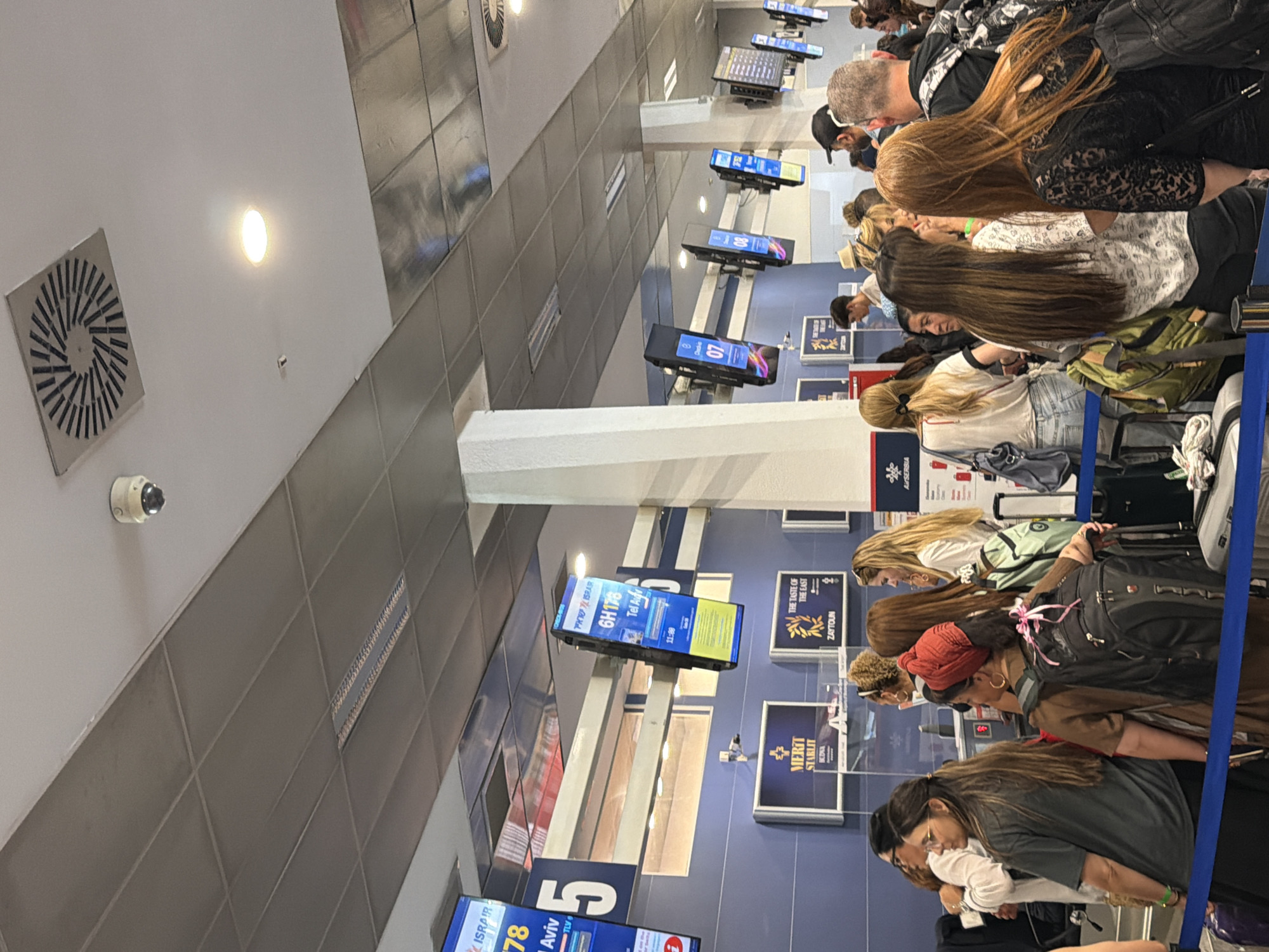 This media file depicts a scene at an airport, likely in Mrčevac, Montenegro, with a long queue of people waiting. The subjects are a diverse group of individuals, primarily seen from behind, with varying hairstyles and clothing. Many are carrying luggage, including suitcases and backpacks, indicating travel. The setting is indoors, characterized by a sterile, tiled wall and a white ceiling with ventilation grates and lighting fixtures. Several digital display screens are visible, showing flight information and advertisements.
The activity taking place is people waiting in line, presumably for check-in, security, or boarding. The overall mood seems to be one of patience, with individuals focused on their phones or looking ahead. There are no clear indicators of time of day or weather.
Notable details include a security camera mounted on the ceiling, various signage with text, and the digital displays. One screen shows "6H178 Tel Aviv" and "11:00," suggesting a flight to Tel Aviv departing at 11:00. Another screen displays "ISRAIR" and "78." Other text includes "THE TASTE OF THE EAST ZAFFOUN" and "MERIT STARLIT BEZENA." The presence of multiple languages on the displays suggests an international airport. The overall impression is a typical airport scene during a busy period.