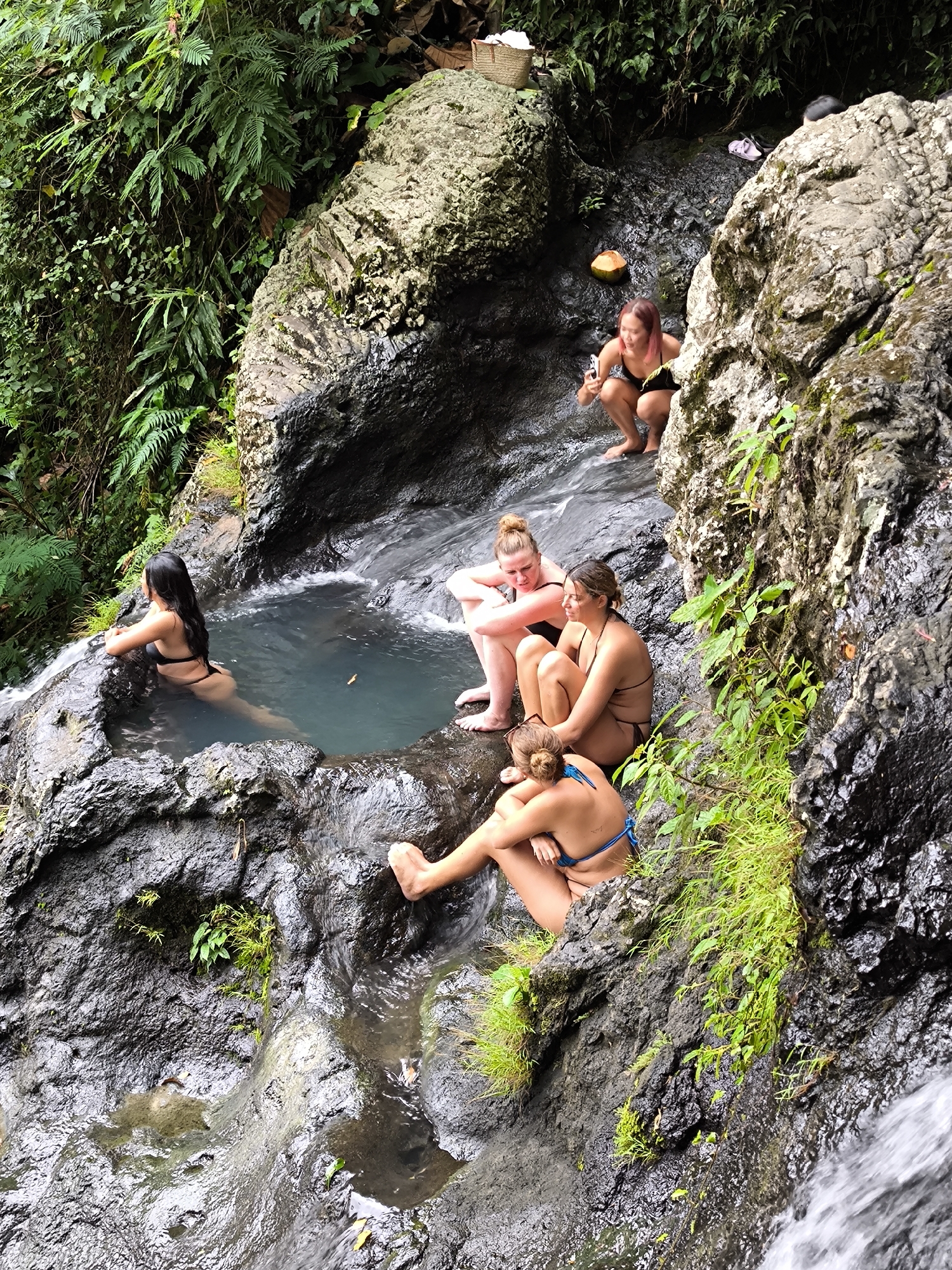The image shows five young women at a rocky waterfall pool in Sidemen, Indonesia. One woman is in the pool, leaning on the edge. Three women sit together on a rock ledge, while another woman is on the rock above them, holding a cell phone and appears to be taking pictures. A coconut rests next to her. The area is lush with green vegetation covering the rocks and surrounding foliage. A small basket is positioned on a rock in the top corner of the image. The water flows down the rocks, creating a waterfall effect. The women are wearing swimwear.
