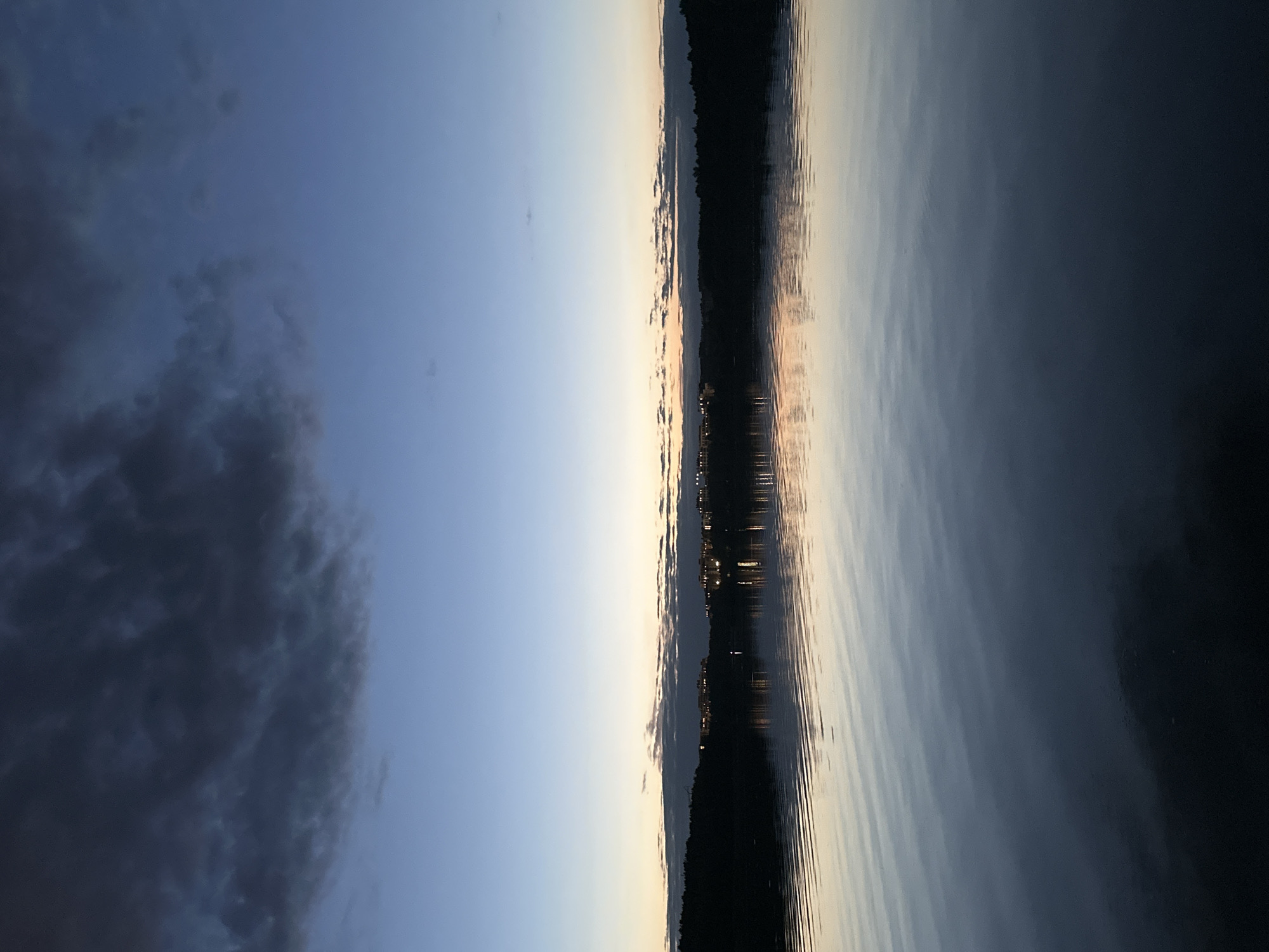 The image depicts a twilight scene over a body of water, likely a lake or river, in Minsk, Belarus. The sky transitions from a deep blue at the top to lighter hues near the horizon, with dark, textured clouds dominating the upper left portion of the frame. Along the distant shoreline, silhouetted trees form a dark band, punctuated by rows of illuminated buildings with lights reflecting on the water's surface. The water itself is calm, with gentle ripples creating subtle patterns that reflect the sky and the lights from the buildings. The overall atmosphere is serene and peaceful, capturing the quiet beauty of a city at dusk.