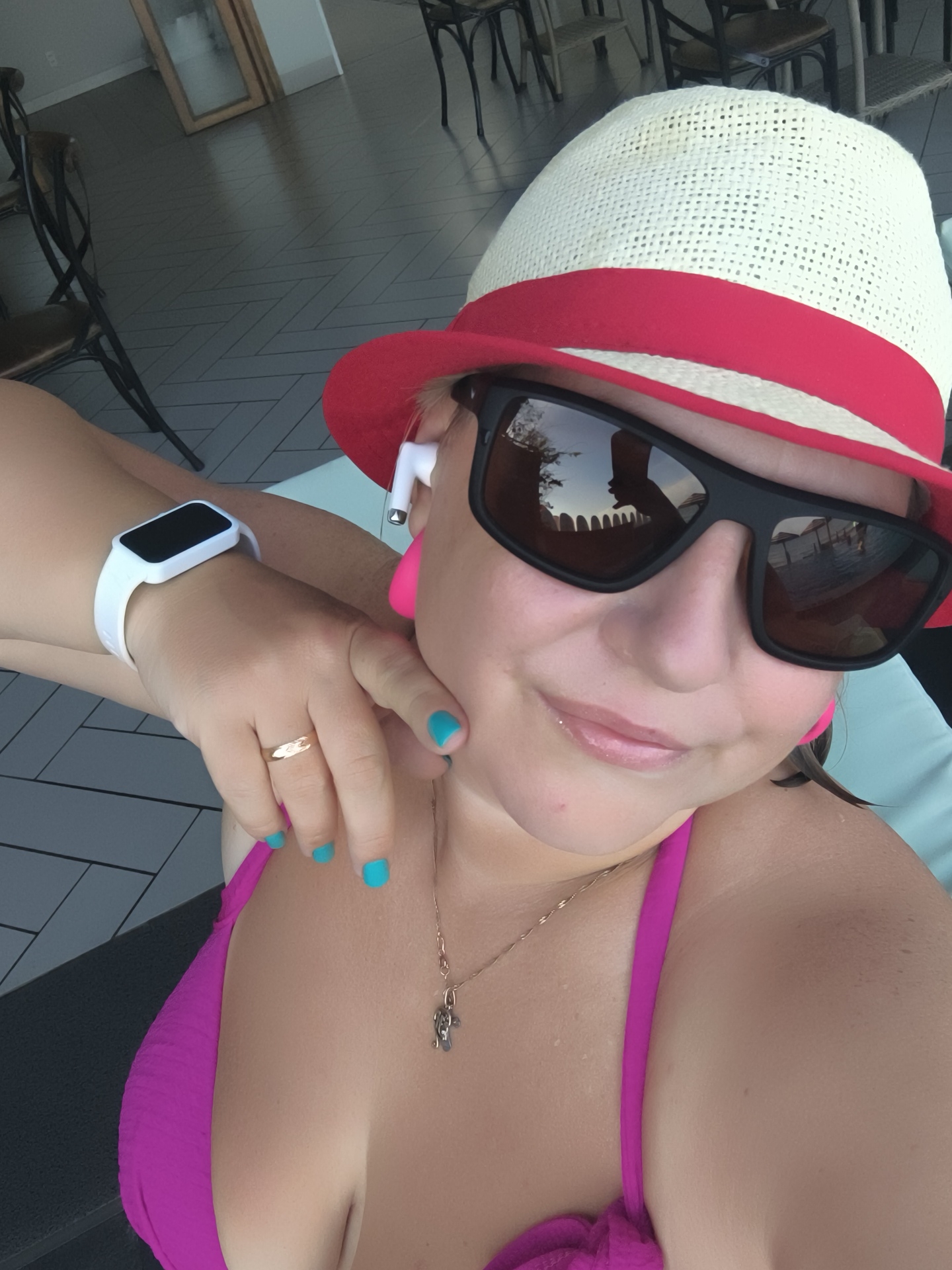 A person wearing a straw hat with a red band, sunglasses, and a pink bikini top is captured in a close-up, selfie-style shot. The individual has bright turquoise nail polish on their fingers and is wearing a white smartwatch on their left wrist and a gold ring on their left ring finger. They are also wearing small white wireless earbuds. The background suggests an outdoor setting with a paved floor and chairs visible. The overall impression is that of a person enjoying a sunny day. The location information provided in the prompt, "Phu Thuy Ward, Vietnam," is not directly discernible from the visual content of the image.