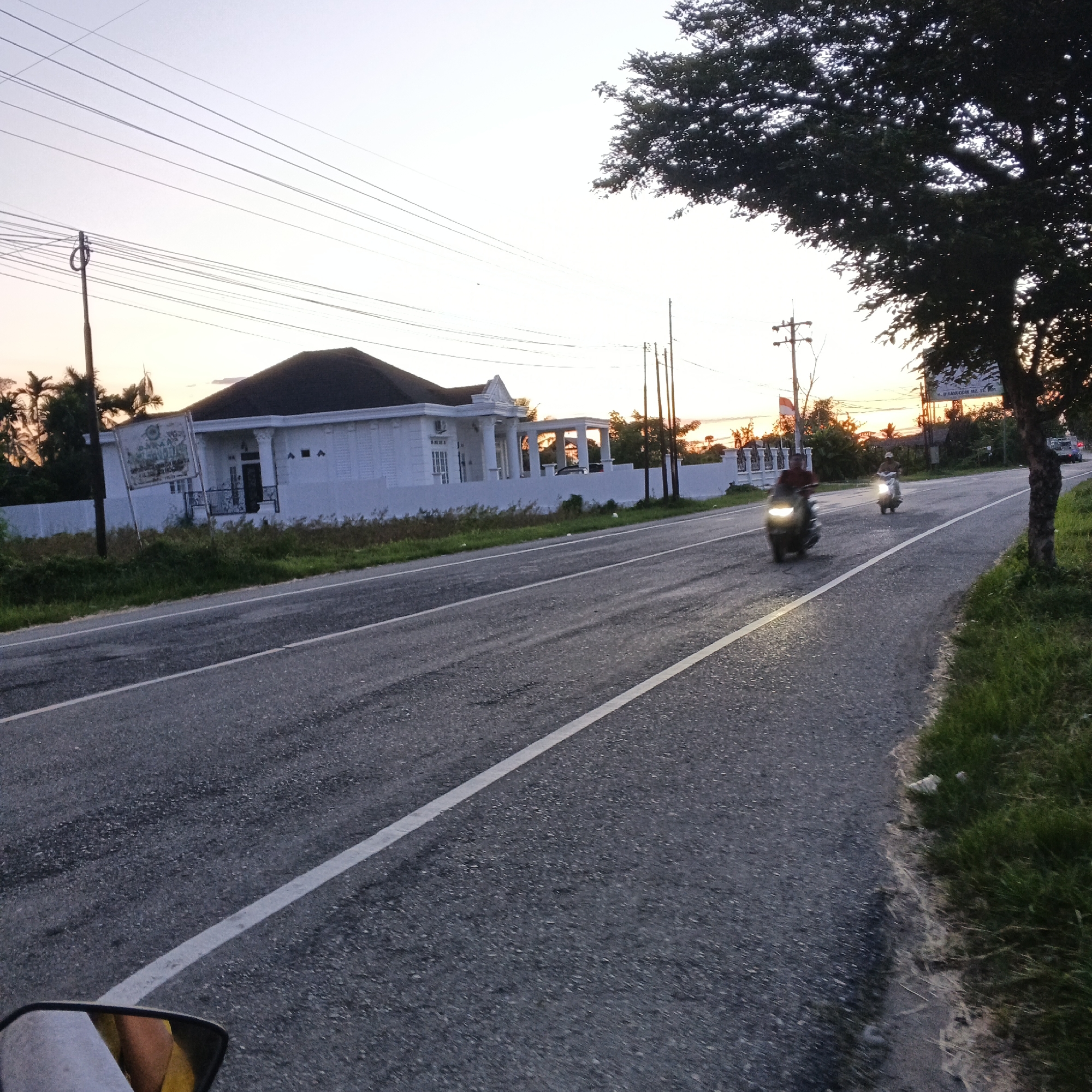 This image captures a street scene in Dayah Baroh, Indonesia, during a sunset. The foreground shows a paved road with a white line marking its center, and a motorcycle's side mirror is visible on the bottom left, reflecting part of a person wearing yellow and white clothing. Two motorcycles are traveling on the road, one with a rider wearing a dark shirt and another with a rider wearing a lighter shirt and a cap. Both motorcycles have their headlights on, illuminating the road ahead.
To the left of the road, a white, modern-style house with a dark roof and classical architectural elements like columns and a portico stands prominently. A sign is mounted on a pole in front of the house, partially obscured by foliage. Tall palm trees are visible behind the house, and a utility pole with multiple wires runs horizontally across the sky.
The right side of the road features a grassy embankment, with a large tree casting a shadow over the pavement. In the distance, the sky is filled with the warm hues of a sunset, with the sun low on the horizon casting an orange glow. The overall environment suggests a residential or semi-rural area. The scene is calm, with the natural light of dusk creating a serene atmosphere.