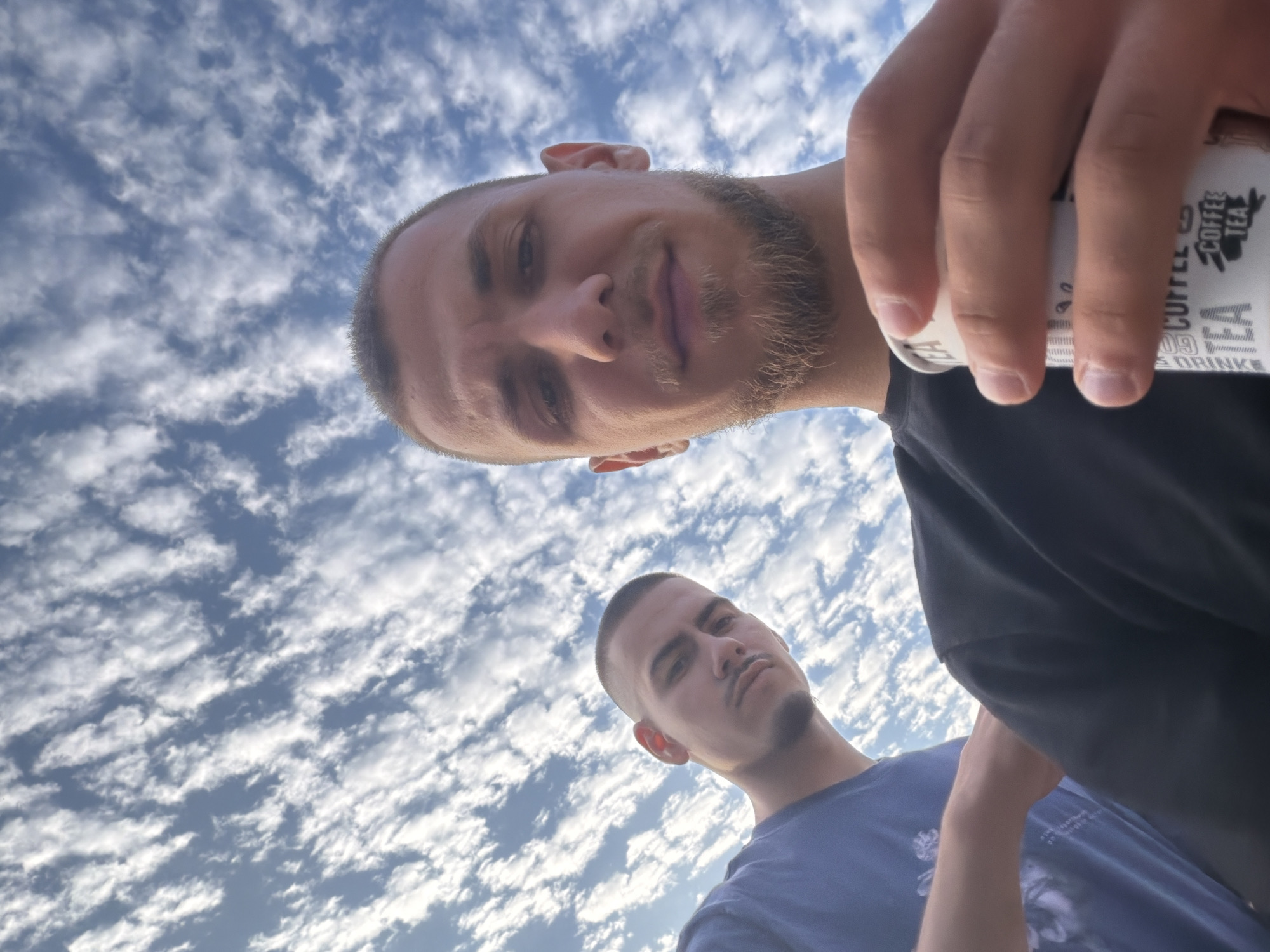 A low-angle selfie captures two young adult males against a bright blue sky filled with numerous white, scattered cumulus or altocumulus clouds. The scene is located in Obnova, Bulgaria.
The man in the immediate foreground, positioned on the right side of the frame, has a light complexion, closely cropped dark hair, and a short, dark beard covering his chin and jawline, extending into a mustache. He wears a plain black crew-neck shirt. His right hand, prominent in the lower right corner, holds a white disposable cup printed with black text, including the words "COFFEE" and "TEA." He looks directly at the camera with a neutral expression.
Behind and to the left of the foreground man, a second young adult male is visible from the chest up. He also has a light complexion, closely cropped dark hair, a thin mustache, and a small soul patch below his lower lip. He wears a solid blue t-shirt. His gaze is directed towards the camera, and his expression appears neutral to slightly serious.