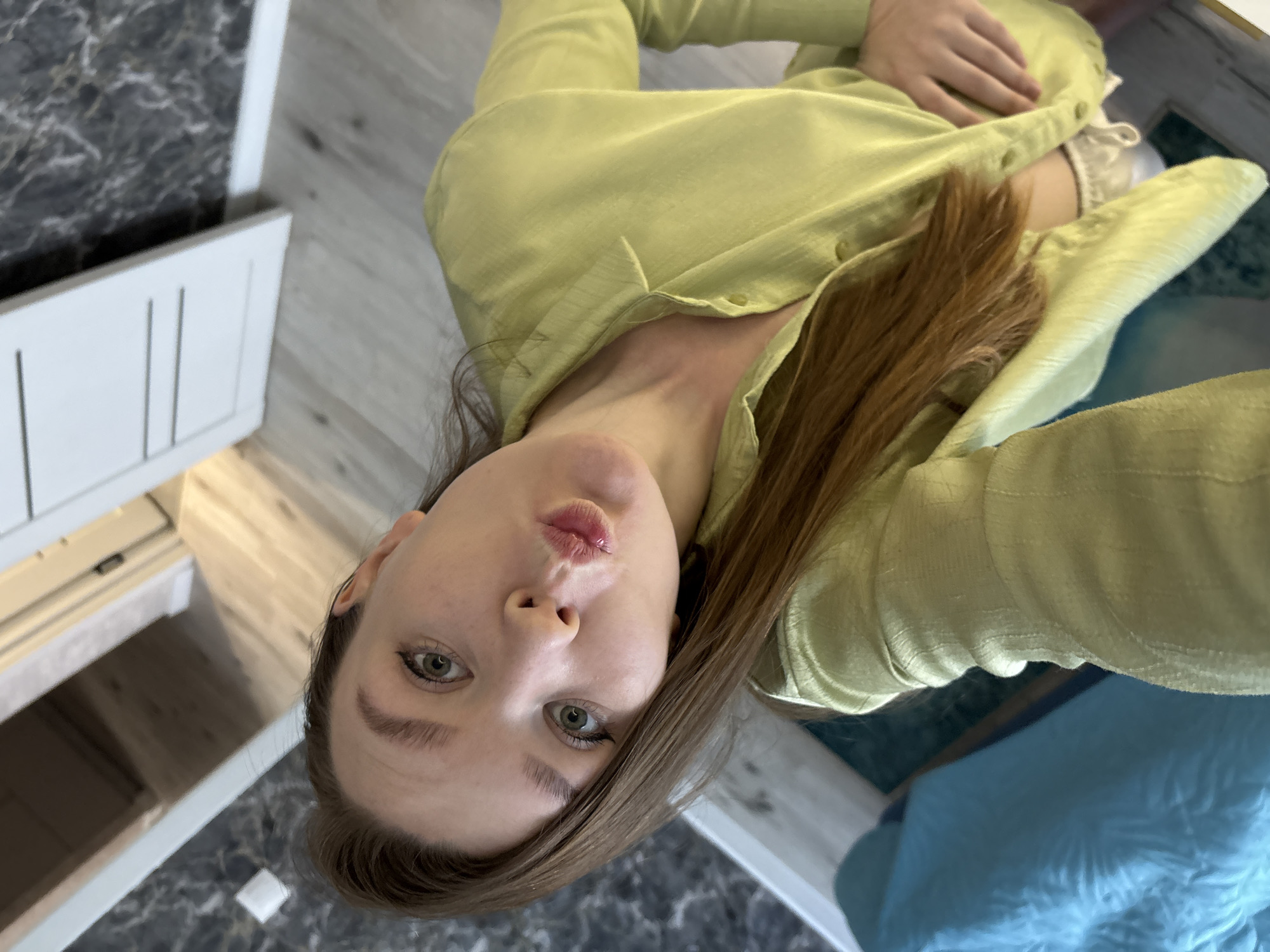 A selfie of a woman with light brown hair wearing a light green button-down shirt. She is looking directly at the camera with pursed lips. The background includes a gray and white tiled floor, a white cabinet or shelf with visible storage space, and a blue cushion or pillow. There is not enough information to determine the location in the city of Izhevsk, Russia.