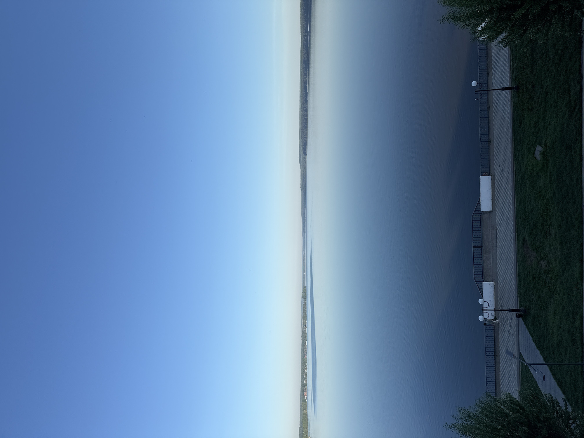 The image shows a vertical view of a river and a portion of the embankment in Votkinsk, Russia. The sky is blue on the left side of the image. The river is on the right and the embankment, with a pathway, lampposts, and grassy areas, occupies the right edge. A line of trees and possibly buildings is visible in the distance, appearing as a thin strip between the sky and the river. There are lampposts with spherical shades are placed along the embankment.