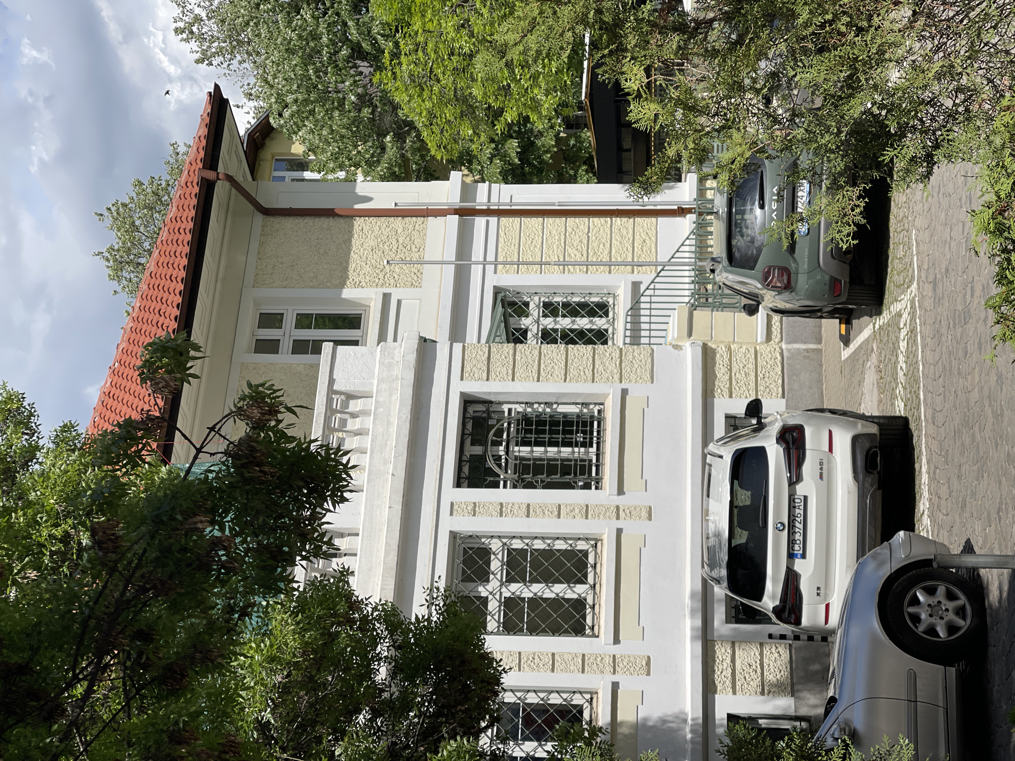 The image captures a three-story building with a red tile roof and a cream-colored facade with white accents. The building features several windows with diamond-patterned metal grilles. A small balcony with a white balustrade is visible on the second floor. A staircase with metal railings leads to an upper level.
Three parked cars are partially visible in front of the building: a green Dacia Duster, a white BMW X3, and a silver Mercedes-Benz.
The setting appears to be an urban environment, with trees partially obscuring the building and cars.
Based on the license plate, the location is Sofia, Bulgaria.