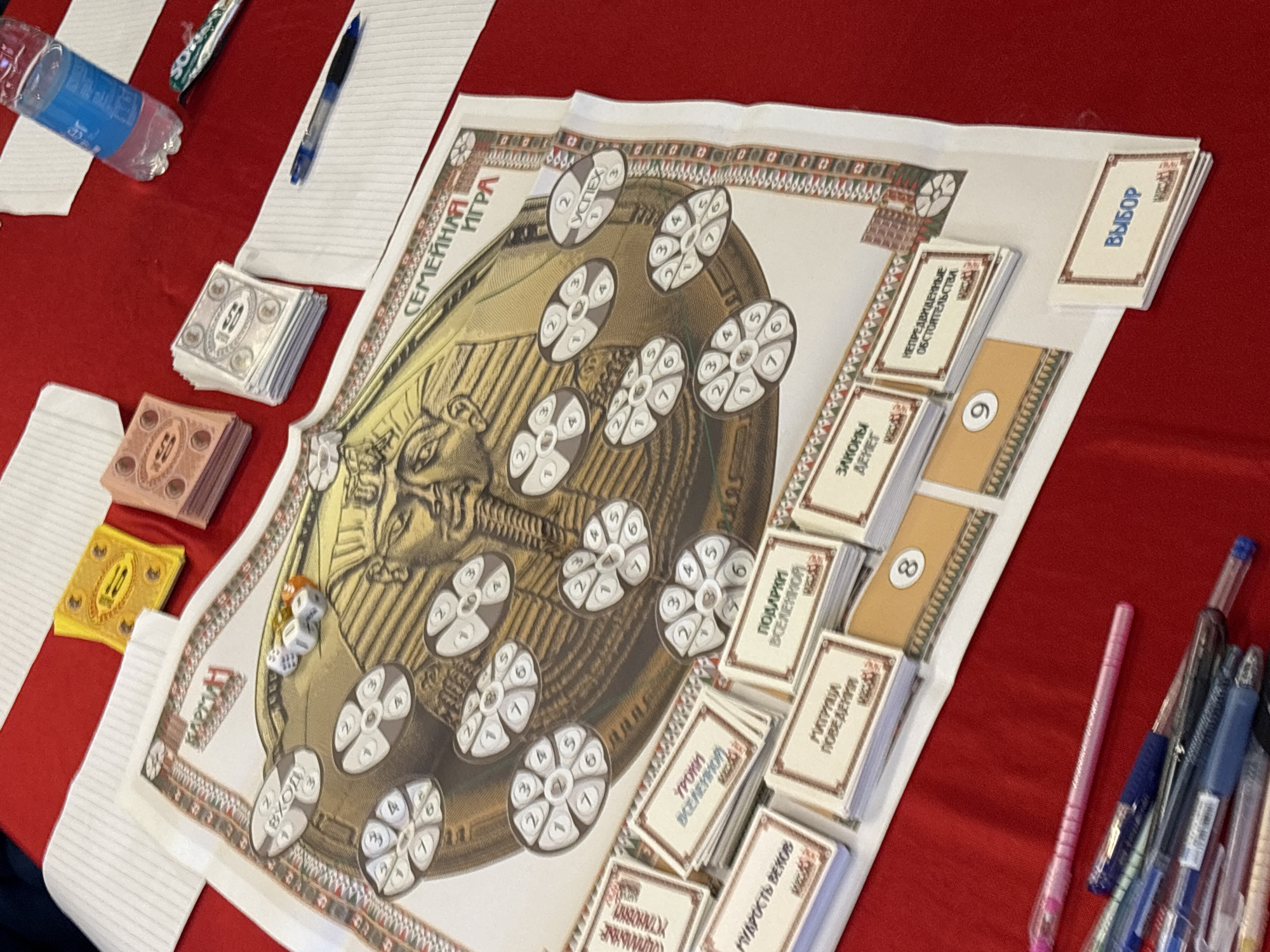 This close-up shot captures a board game laid out on a red tablecloth, possibly in Kuta Selatan, Indonesia. The main game board features a large, golden stylized head resembling an ancient Egyptian pharaoh or deity, surrounded by numerous circular tokens. Most tokens display numbers from 1 to 7, arranged like segments of a wheel. One token prominently displays the word "УСПЕХ" (Success). The title "СЕМЕЙНАЯ ИГРА" (Family Game) is visible on the board's border.
Various stacks of game cards are neatly arranged around the board. Text on these cards includes "ЗАКОНЫ ДЕНЕГ" (Laws of Money), "НЕПРЕДВИДЕННЫЕ ОБСТОЯТЕЛЬСТВА" (Unforeseen Circumstances), "ПОДАРКИ ВСЕЛЕННОЙ" (Gifts of the Universe), "УРОКИ ВСЕЛЕННОЙ" (Lessons of the Universe), "МУДРОСТЬ ВЕКОВ" (Wisdom of Ages), "МАТРИЦА ПОВЕДЕНИЯ" (Matrix of Behavior), and "ВЫБОР" (Choice). There are also card stacks labeled with numbers "8" and "9".
Stacks of game money are also present, with some showing "50" on silver-colored notes and "10" on yellow notes. Two white dice with black pips are seen on the board. Other items on the red table include a blue water bottle, several pens (including a pink one), and sheets of white paper, suggesting a game in progress or about to begin.