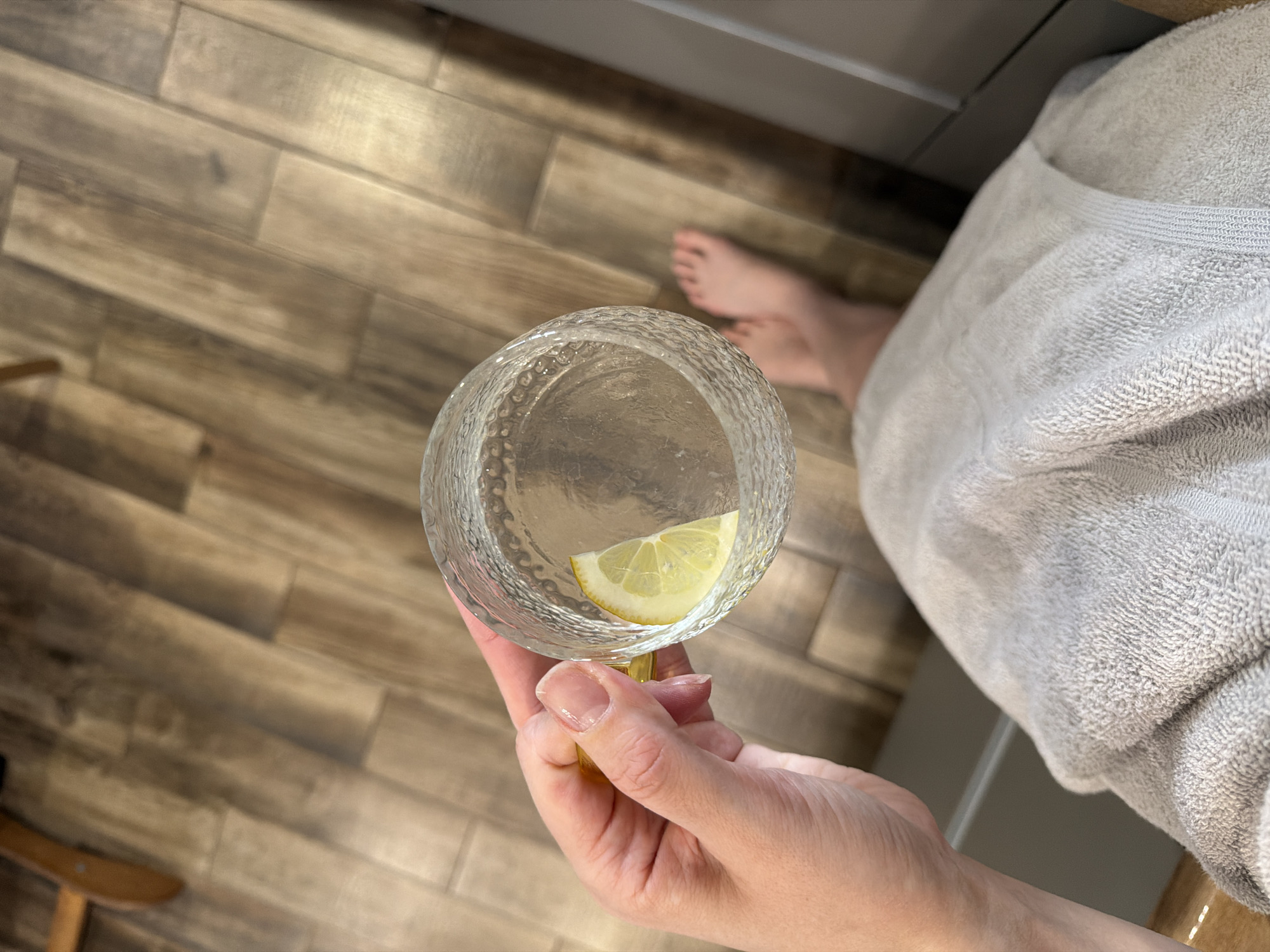A first-person overhead shot shows a hand holding a textured glass containing water and a lemon slice. The hand has polished fingernails. In the background are bare feet, a gray towel or robe, and light brown wood-patterned tiles. The location is not specified, and there is no information to indicate it is in Chaikovsky, Russia.