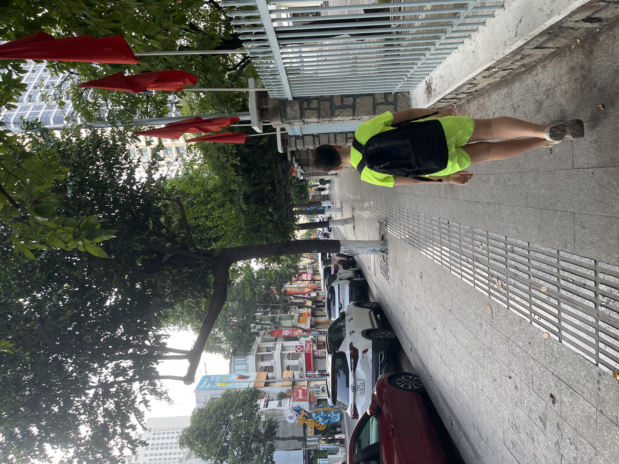 The image depicts a street scene in Nha Trang, Vietnam. A young person wearing a neon green t-shirt and shorts, with a black backpack, walks away from the camera on a sidewalk. The sidewalk has tactile paving for the visually impaired. To the right of the sidewalk is a stone wall with a light blue metal fence. Red flags are visible on poles behind the fence. A row of parked cars, including a white Kia, lines the street on the left. Beyond the cars, buildings with shops and balconies are visible. Trees line the street, casting shadows on the sidewalk. The sky is bright, suggesting it is daytime.