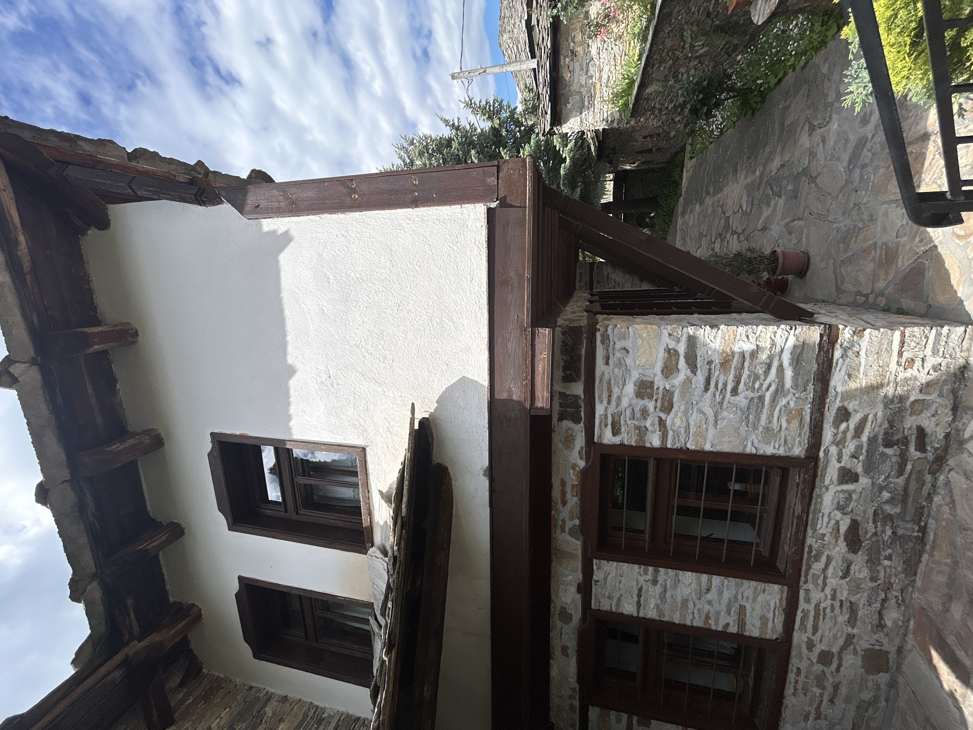 The image shows a building in Dolen, Bulgaria, with a combination of white stucco and stone construction. The building has dark wood trim around the windows and at the corners. Windows are visible on both the stucco and stone sections. A stone pathway is nearby. The sky is visible with clouds.