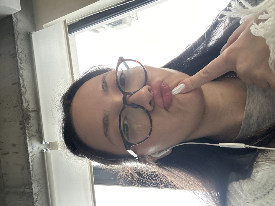 An indoor portrait displays a female individual, shown from the upper chest upward. She has dark hair, dark-framed eyeglasses with tortoiseshell patterns, and a white wired earphone inserted into her left ear. Her lips are pursed in a prominent "duck face" expression, and her right index finger, adorned with a pearlescent white manicured nail, is positioned on her chin below her lower lip. She is wearing a light-colored, possibly cream or white, chunky knit garment over a visible grey top. The background consists of a rough concrete ceiling beam overhead, featuring a visible mounting bracket and what appears to be a light fixture. Below the beam, a partially lowered white roller blind is visible. To her right (viewer's left), a large window lets in bright, diffuse light, through which faint outlines of exterior structures are indistinctly visible. The scene is situated in an interior location within Yerevan, Armenia.