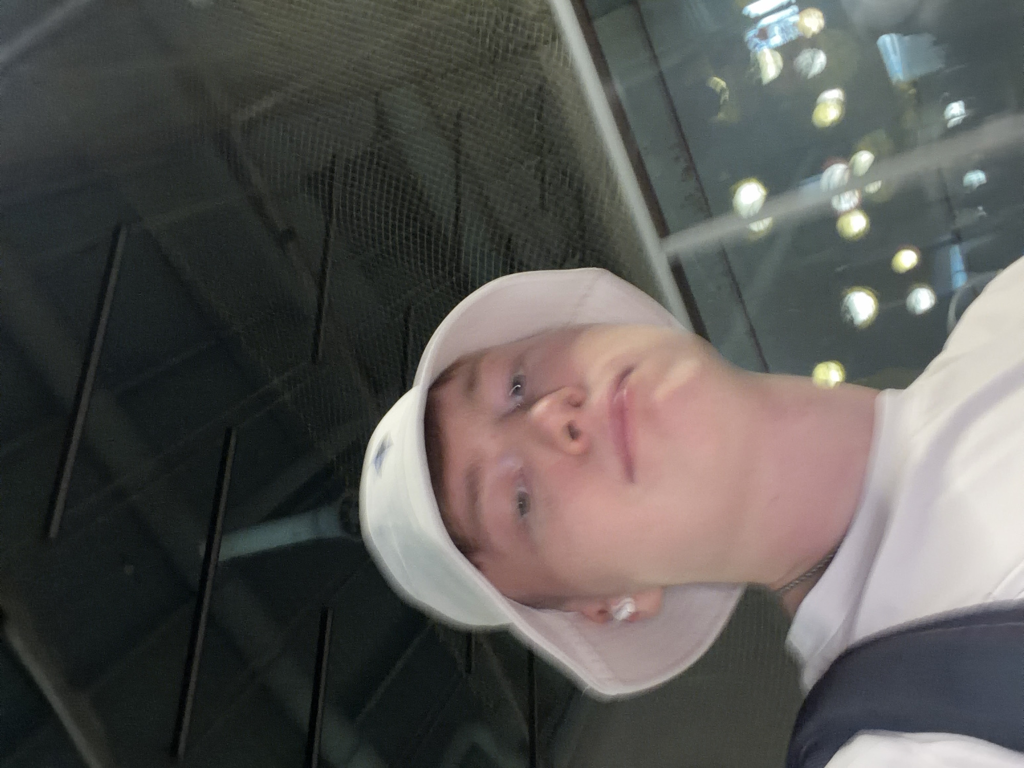 The image is a selfie taken from a low angle, showing a person wearing a white bucket hat and a white t-shirt. The person has fair skin and light-colored hair, and is wearing a white wireless earbud in their left ear. They are also wearing a silver chain necklace. The background is dark, with some out-of-focus lights visible, suggesting an indoor or nighttime setting. The ceiling structure is visible above the person's head, appearing to be made of dark material with linear elements. There is a mesh-like material or netting also visible in the upper part of the background. The overall lighting is dim, with a slight yellowish cast from the lights in the background. The location is specified as Kyiv, Ukraine, but this information is not visually deducible from the image itself.