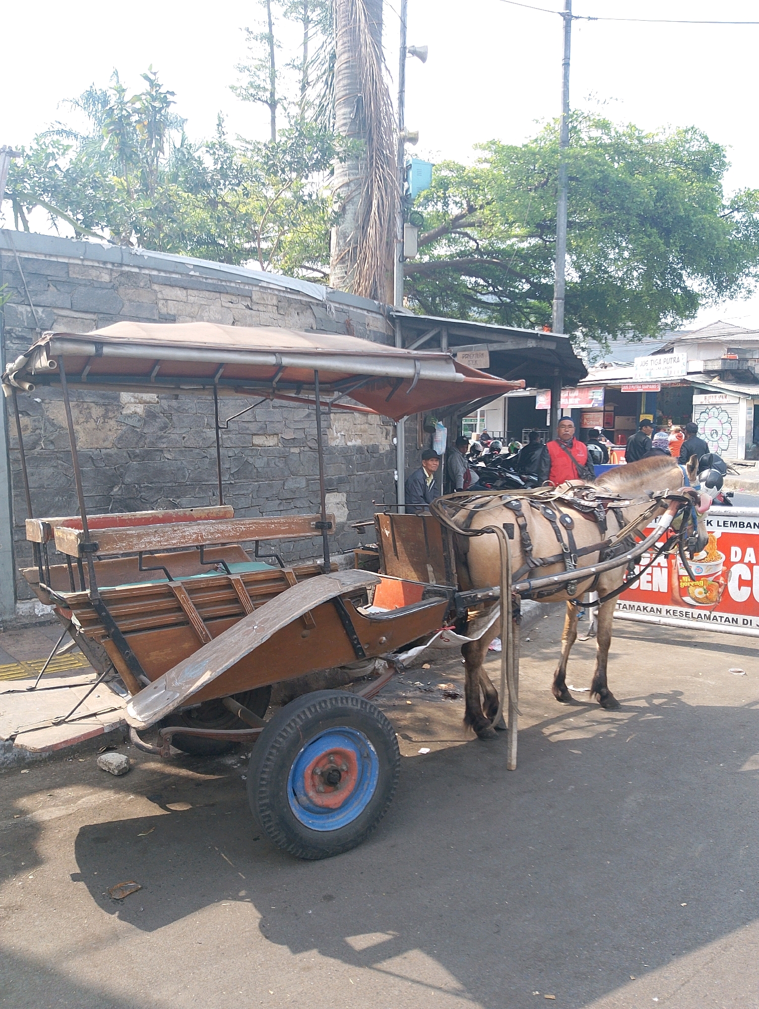 In Lembang, Indonesia, a horse-drawn carriage, likely a delman or delman kuda, is parked on a paved street beside a low stone wall. The carriage, made of wood with a canvas roof, features two rows of wooden seats. A light brown horse is harnessed to the front of the carriage, standing patiently. Several people are visible in the background, some near a building with a sign that reads "JUS TIGA PUTRA," suggesting a juice vendor. Another person in a red vest is standing near the horse and carriage. The environment appears to be an urban or semi-urban setting with trees and a bright, overcast sky. The scene captures a moment of everyday life, possibly waiting for passengers or taking a break from service.