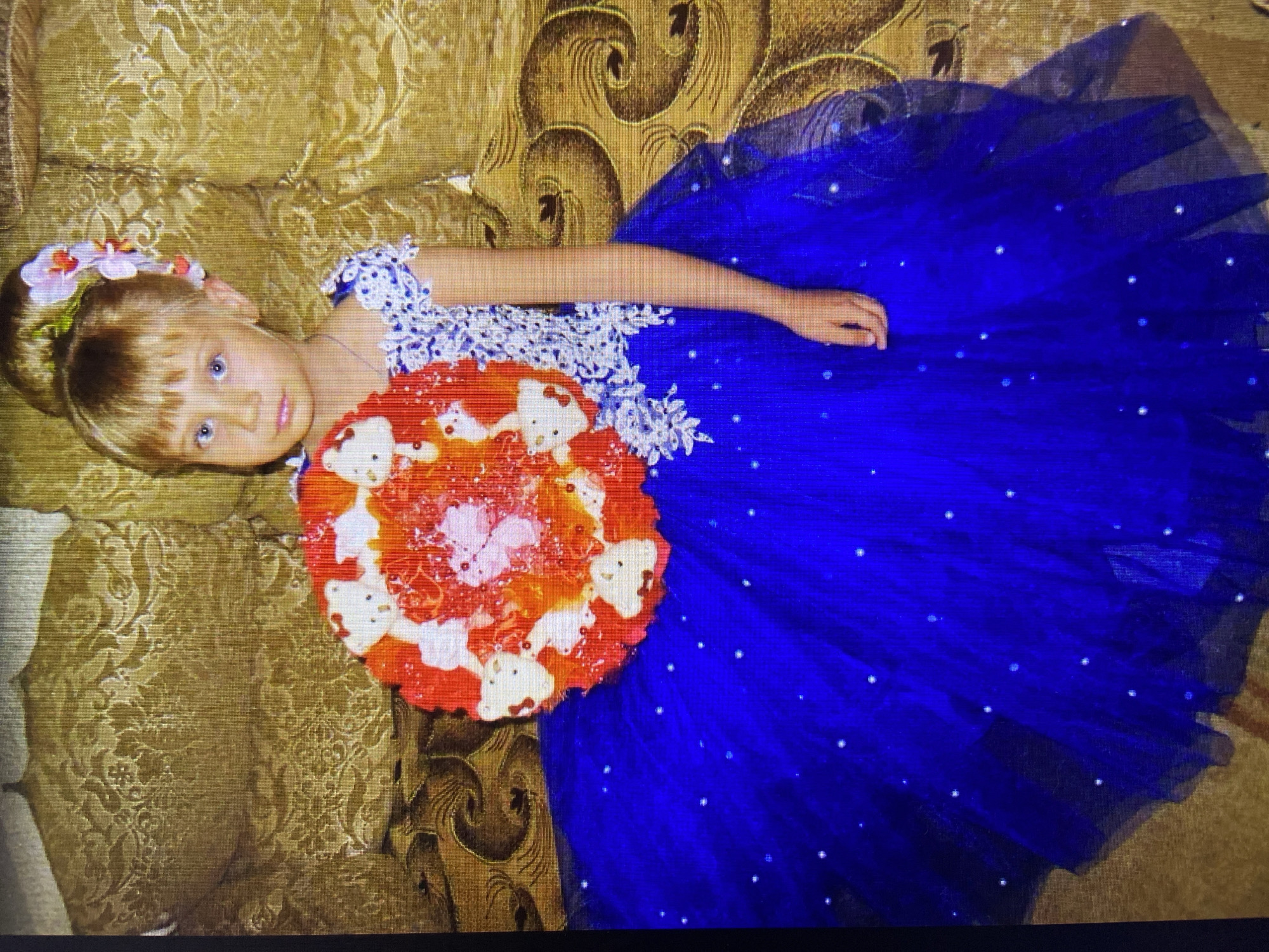 A young girl with blonde hair styled up with pink orchids stands in front of a gold upholstered couch. She is wearing a blue, floor-length gown adorned with white sparkly dots. She holds a round bouquet consisting of red and white decorations, possibly candy or flowers. The girl is looking directly at the camera with a neutral expression. The background shows the ornate pattern of the couch. The image quality is poor, with visible pixelation and color distortion, suggesting it may be a low-resolution or scanned photograph. There is no information available to determine the specific location in Stavropol, Russia.