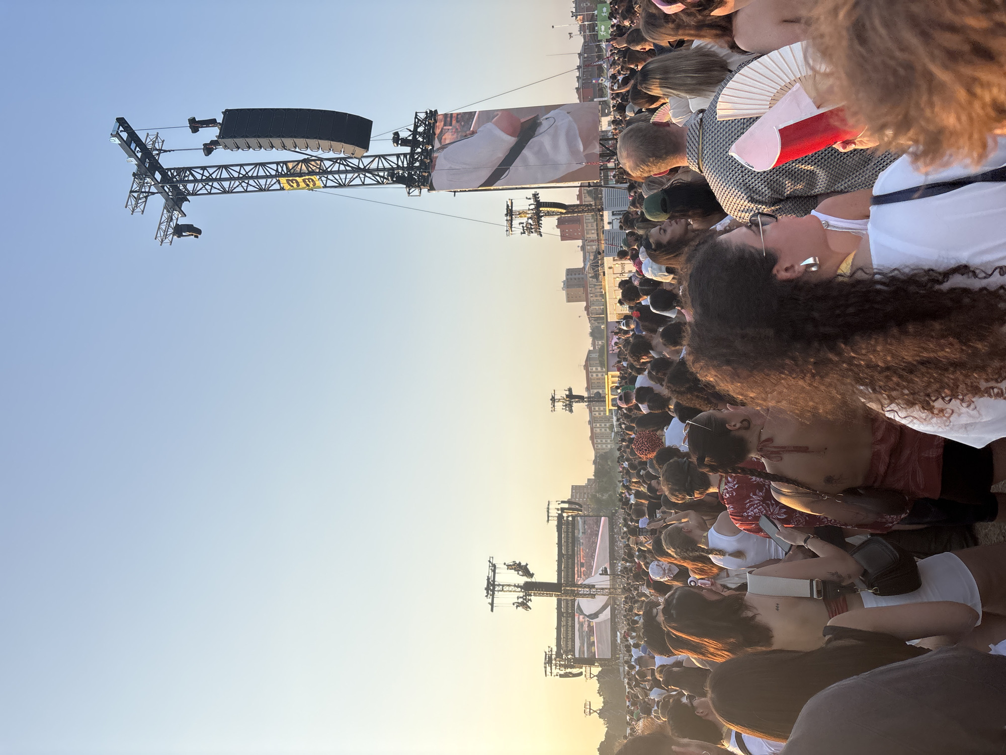 This image depicts a large outdoor music festival with a massive crowd gathered under a clear, pale blue sky. The sun is low in the sky, casting a warm, golden light, suggesting it is late afternoon or early evening. In the background, several large stage structures are visible, equipped with large screens displaying imagery, and arrays of speakers. These structures are tall, industrial-looking metal frames with multiple crossbeams and support elements. One prominent screen shows a close-up of a person wearing a white t-shirt and a black strap, possibly a musician or performer. In the foreground and midground, a dense crowd of people of diverse ethnicities and ages are present. They are dressed in casual, summer attire, including t-shirts, tank tops, and shorts. Many individuals are holding up their phones, likely capturing the event. Some people are holding fans, such as the white and pink fan visible in the right foreground, to combat the warmth. The overall atmosphere appears to be one of excitement and shared experience. The presence of large-scale concert infrastructure and the urban skyline in the distance suggests this is an event held within a city. Given the context of the concert and the architectural style of the buildings in the background, it is plausible that this event is taking place at the Ippodromo Snai in Milan, Italy, a known venue for large concerts and festivals.