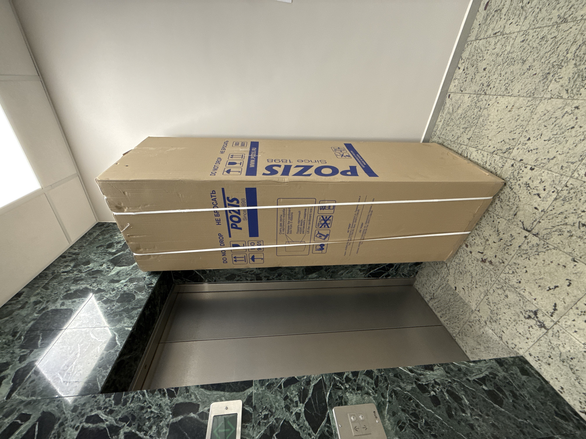 This image depicts a large cardboard box situated in an elevator lobby or hallway, likely in a commercial or residential building in Moscow, Russia. The floor is distinctly patterned, with dark green marble-like tiles on the left and lighter speckled granite-like tiles on the right, meeting at a diagonal. The walls are a plain, light beige color.
The primary subject is a long, rectangular cardboard box, secured with white plastic straps. It is positioned near an open elevator with stainless steel doors. The box is prominently branded "POZIS" and states "Since 1898," along with "WWW.POZIS.RU," "ISO 9001," and "MADE IN RUSSIA" (ИЗГОТОВЛЕНО В РОССИИ). Important handling instructions and warnings are visible: "DO NOT DROP" in both English and Russian (НЕ БРОСАТЬ), symbols for "Lift up" and fragility. Additional Russian text provides unpacking instructions: "Снять две ленты с картонного короба" (Release the cardboard from 2 ribbons) and "Поднять картонный короб вверх, распаковать изделие" (Lift the cardboard box up, unpack the product).
An elevator control panel with "OTIS" branding and up/down arrows is visible on the dark green tiled floor. The box's size suggests it contains a large appliance. The scene implies that the box is either waiting to be loaded into the elevator or has just been unloaded, indicating an activity of moving goods within the building. The indoor setting is well-lit by artificial lighting.