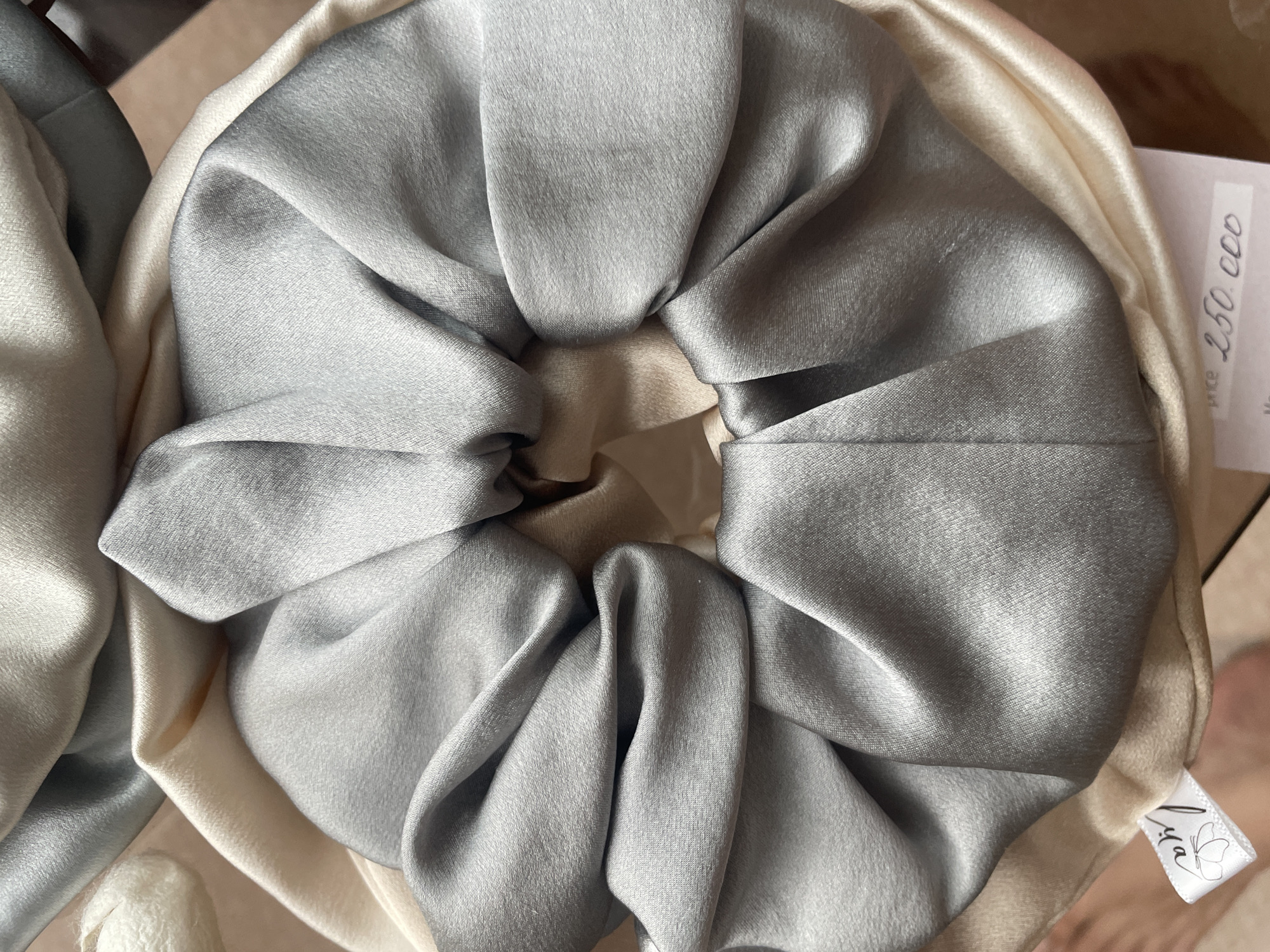 The image shows a close-up of two scrunchies. One is a shiny, light gray color, and the other is a lighter, beige or cream color. The scrunchies appear to be made of silk or a similar fabric. A price tag is visible with handwritten text (including "250.000"). A small tag with a logo and the word "Lya" is also visible on the beige scrunchie. The background is out of focus, but suggests a neutral-colored surface. The image appears to have been taken indoors. Bac Nha Trang Ward, Vietnam is mentioned, but the image contains no context that specifies this location. Therefore it cannot be included in the description.