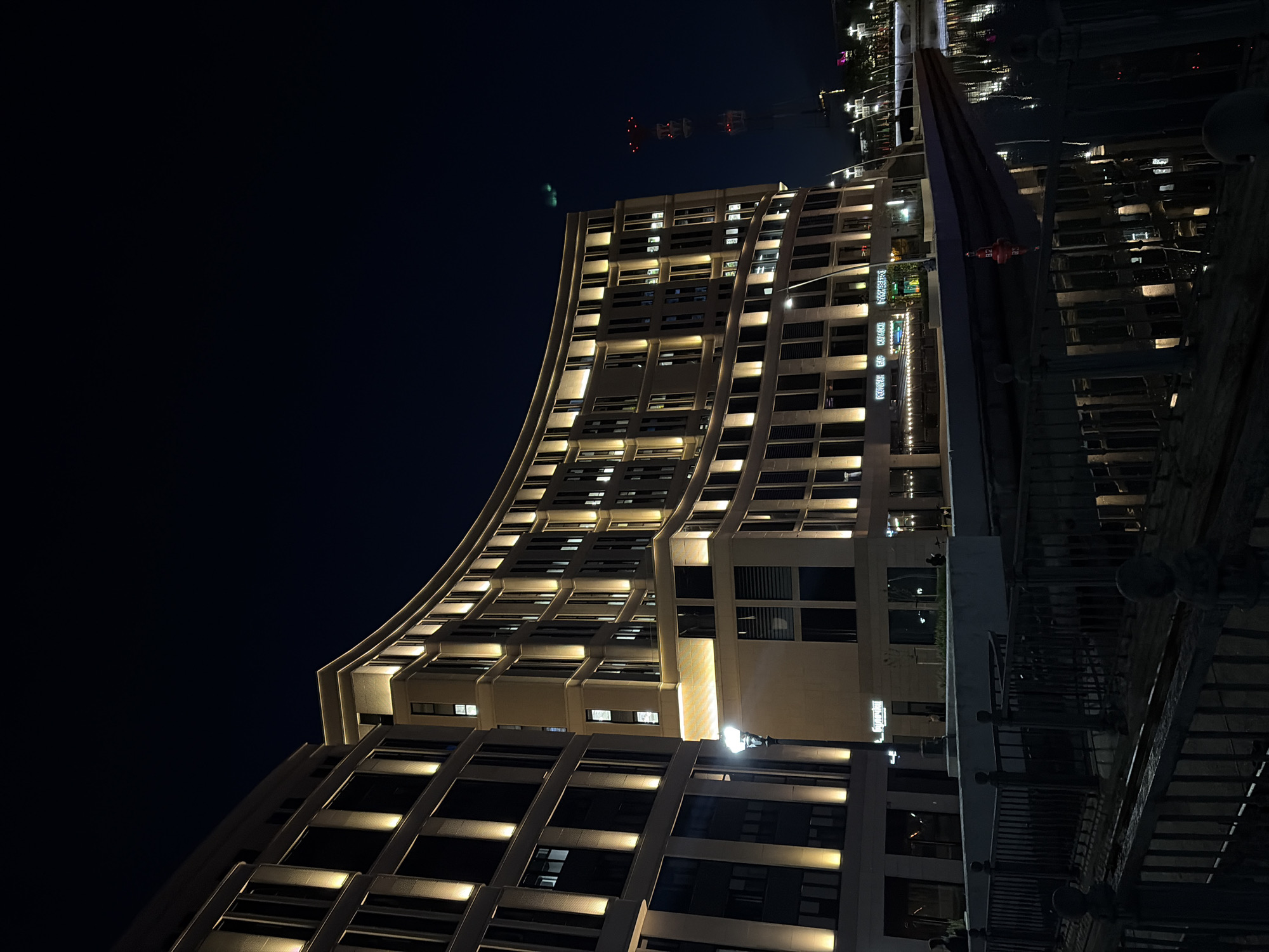 A low-angle, night-time shot features a modern architectural building with illuminated windows. The building appears to have a curved facade, rising from the lower right corner diagonally into the frame. Adjacent to the building, what seems to be a waterfront pier or walkway is visible, with a metal railing running along its edge. The sky above is dark, and a few distant lights or structures can be seen in the background. The city is unknown.