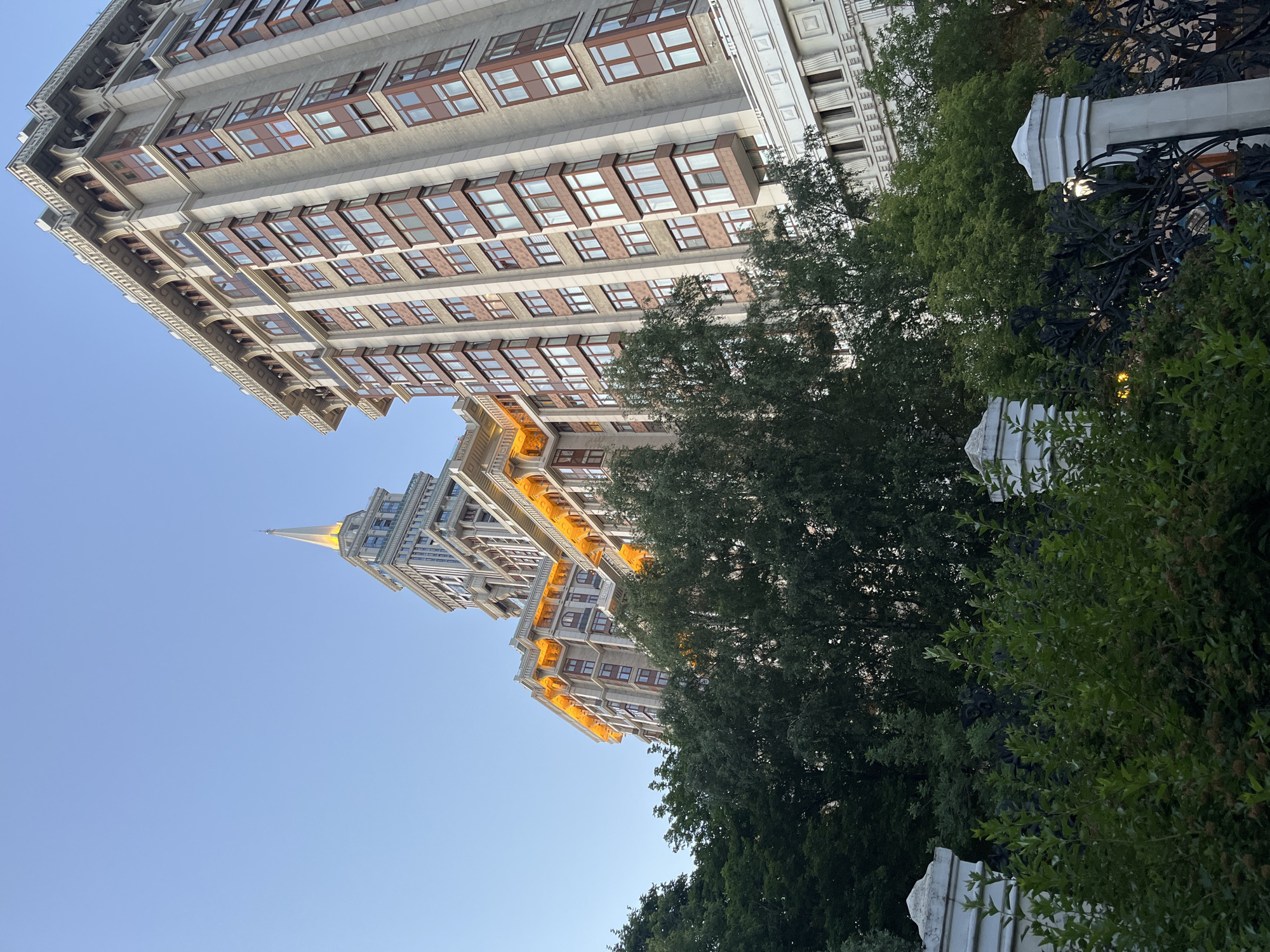 This is a low-angle, exterior, eye-level, full, medium shot of the Stalinist skyscraper located in Khimki, Russia, on a clear day. The skyscraper is made of concrete and stone, with multiple floors of windows and decorative elements. The spire of the building is illuminated with a warm yellow light, as are the upper floors of the adjacent building. Lush green trees and bushes surround the base of the building. The foreground features a white fence with ornate black ironwork, partially obscured by the foliage. There are no people visible in the image. The overall impression is one of grandeur and architectural splendor.