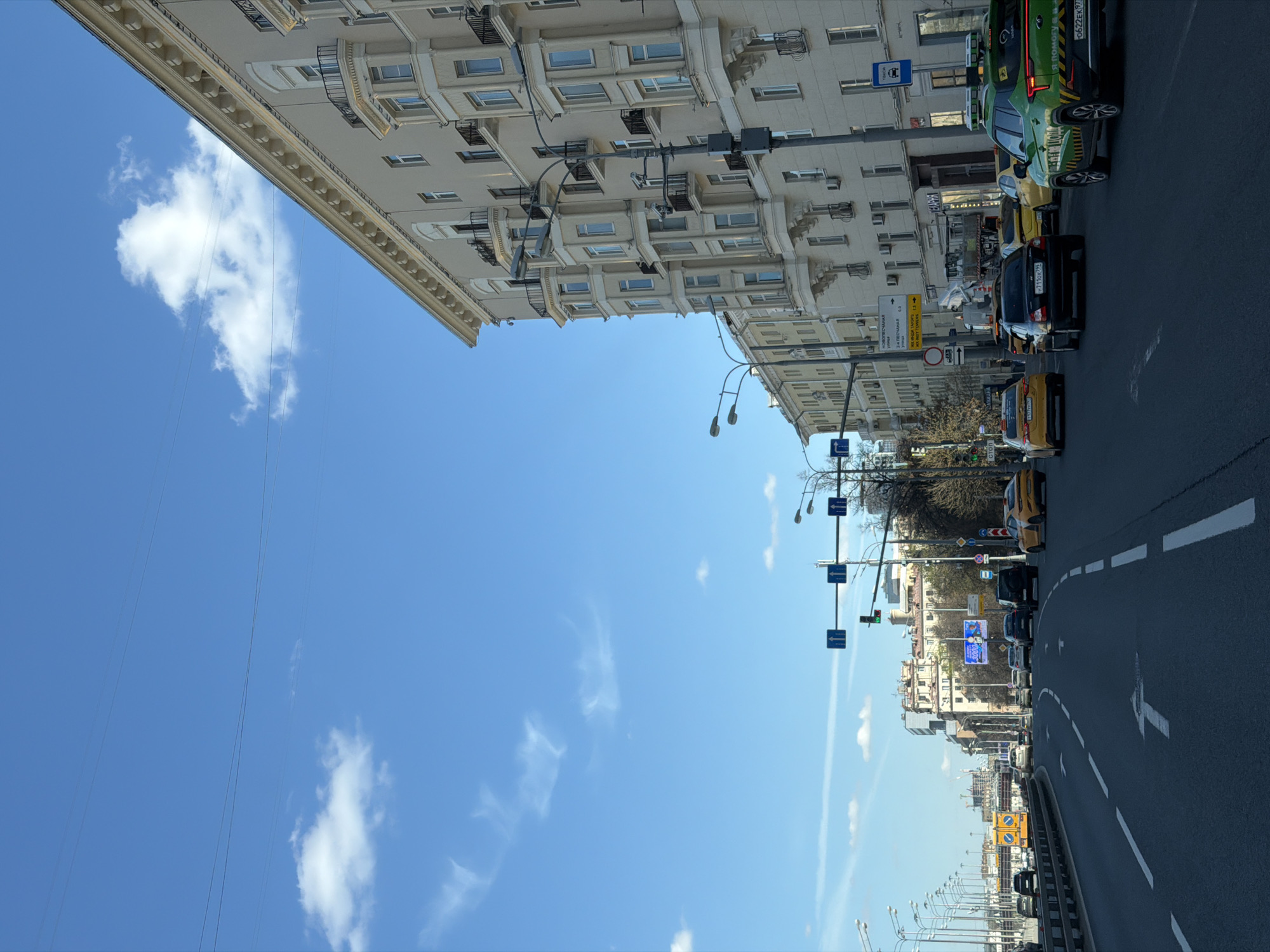 A vertical shot depicts a city street in Khimki, Russia, under a bright blue sky with scattered clouds. A large building lines one side of the street, its facade detailed with numerous windows and decorative elements. Traffic flows on the asphalt road, including cars and taxis, some of which are distinctively colored. Streetlights and traffic signs are positioned along the road, and the urban landscape extends into the distance.