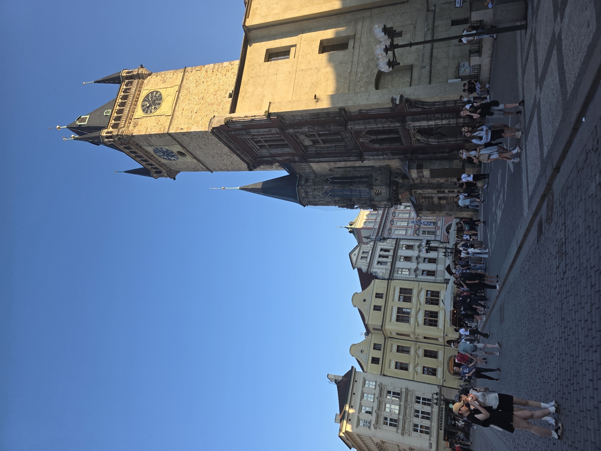This image captures a vibrant street scene dominated by historic European architecture, strongly suggesting Prague's Old Town Square.
**Subjects:** The central subject is a towering, ornate stone clock tower, likely the Old Town Hall Tower, featuring at least two distinct clock faces. Surrounding it are numerous multi-story historic buildings with varying architectural styles, rooflines, and windows. Many people, mostly young adults, populate the cobblestone street and sidewalks, appearing in groups or individually. A street lamp with three spherical lights is also visible.
**Activity or event taking place:** People are engaged in typical street activities: walking, standing, and conversing. There is no specific organized event, but rather the general hustle and bustle of a popular urban area.
**Notable details:** The time of day appears to be late afternoon or early evening, indicated by the warm, directional sunlight casting shadows on the buildings and street, under a clear, bright blue sky. The prominent clock tower and surrounding architecture are strong cues for Prague.
**Visible text:** The word "STARBUCKS" is clearly visible on a storefront sign on one of the buildings. Another building displays the word "CAFE."