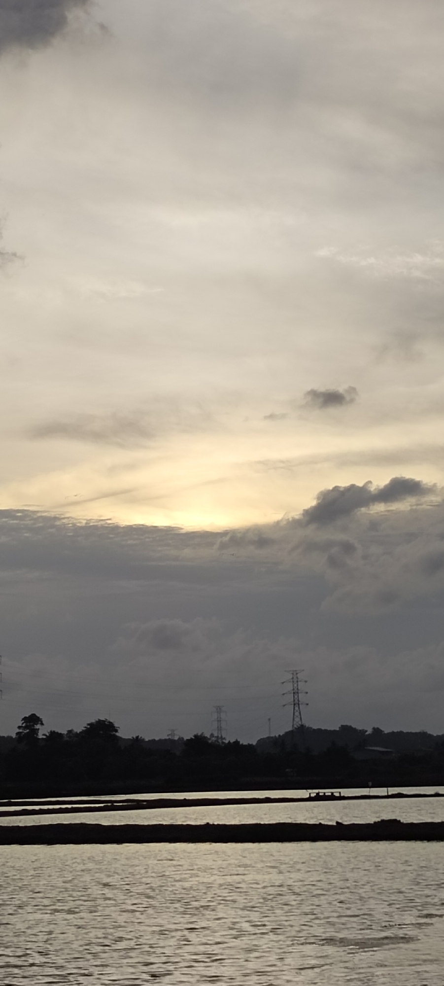The image depicts a landscape in Lhokseumawe, Indonesia, at dusk or dawn. The foreground features a body of water, likely rice paddies, with land dividing sections. The reflection of the sky is visible on the water's surface. The middle ground contains silhouetted trees and shrubbery, interspersed with power pylons stretching across the horizon. The sky is predominantly overcast with clouds obscuring most of the sun except for a narrow strip. The overall tone is muted, with limited color. No people or animals are visible in the scene.