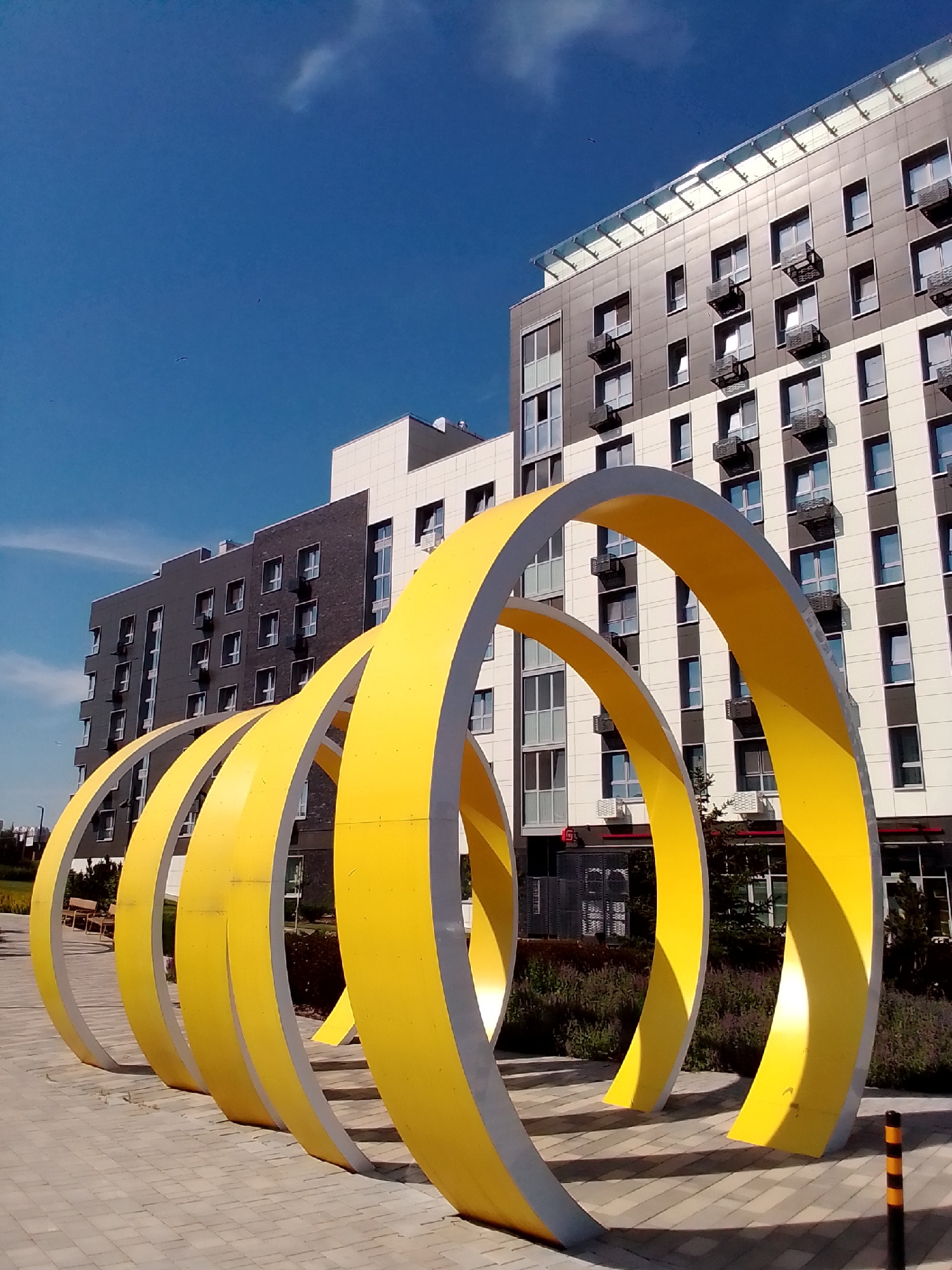 A row of large, bright yellow, curved sculptures stands in front of two modern, multi-story buildings under a clear blue sky. The sculptures are made of thick, curved metal painted yellow on the inside and a light gray on the outside. They are arranged in a receding line, creating a sense of depth. The buildings behind the sculptures have a mix of dark gray and light-colored facades with numerous windows and air conditioning units. The ground in front of the sculptures is paved with light gray rectangular tiles. In the background, a patch of dark green bushes and some smaller trees are visible. To the far left, a wooden bench can be seen. A black and yellow striped bollard stands to the right of the frame. The scene appears to be an urban outdoor public space.