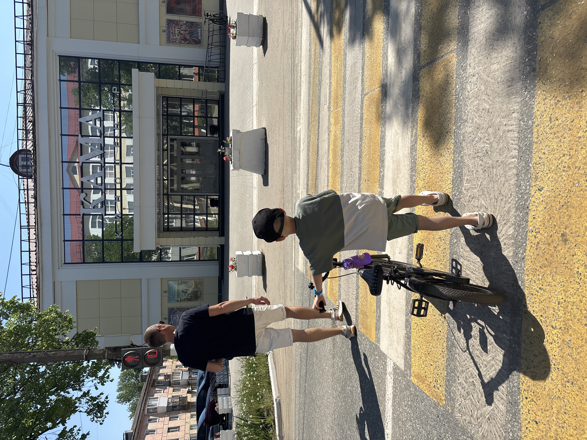 A sunny day in Chaikovsky, Russia, presents a scene at a pedestrian crossing. The building directly in the background is the "Sayany" cinema. A man in a black shirt and white shorts stands on the left side of the crosswalk. A child in a green and white shirt, black cap, and sneakers stands in the center, holding a black bicycle with a purple water bottle. The crosswalk consists of yellow and white stripes. The building behind the crosswalk has large glass windows and doors. The sign on the building reads "SAYANY". Traffic light is on the pole at the street.