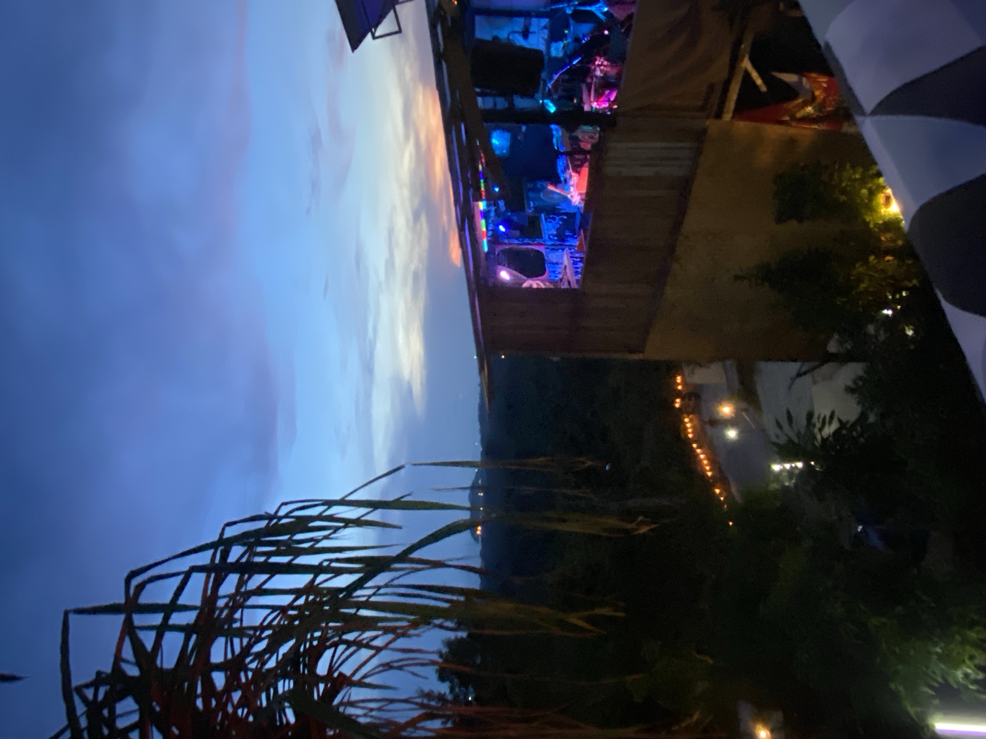 The image depicts a nighttime scene in Baan Don Sai, Thailand. In the foreground, tall reeds or grass stalks are silhouetted against a blue and pink-hued sky. Midground shows a dark, forested hillside. In the background, a raised, wooden structure with an illuminated interior is visible. The interior is lit with vibrant blue and purple lights, suggesting a gathering or performance. A checkerboard pattern is present, partially obscuring a tree, on the right edge of the frame. Dim lights are visible along the hillside, potentially indicating a road or path.
