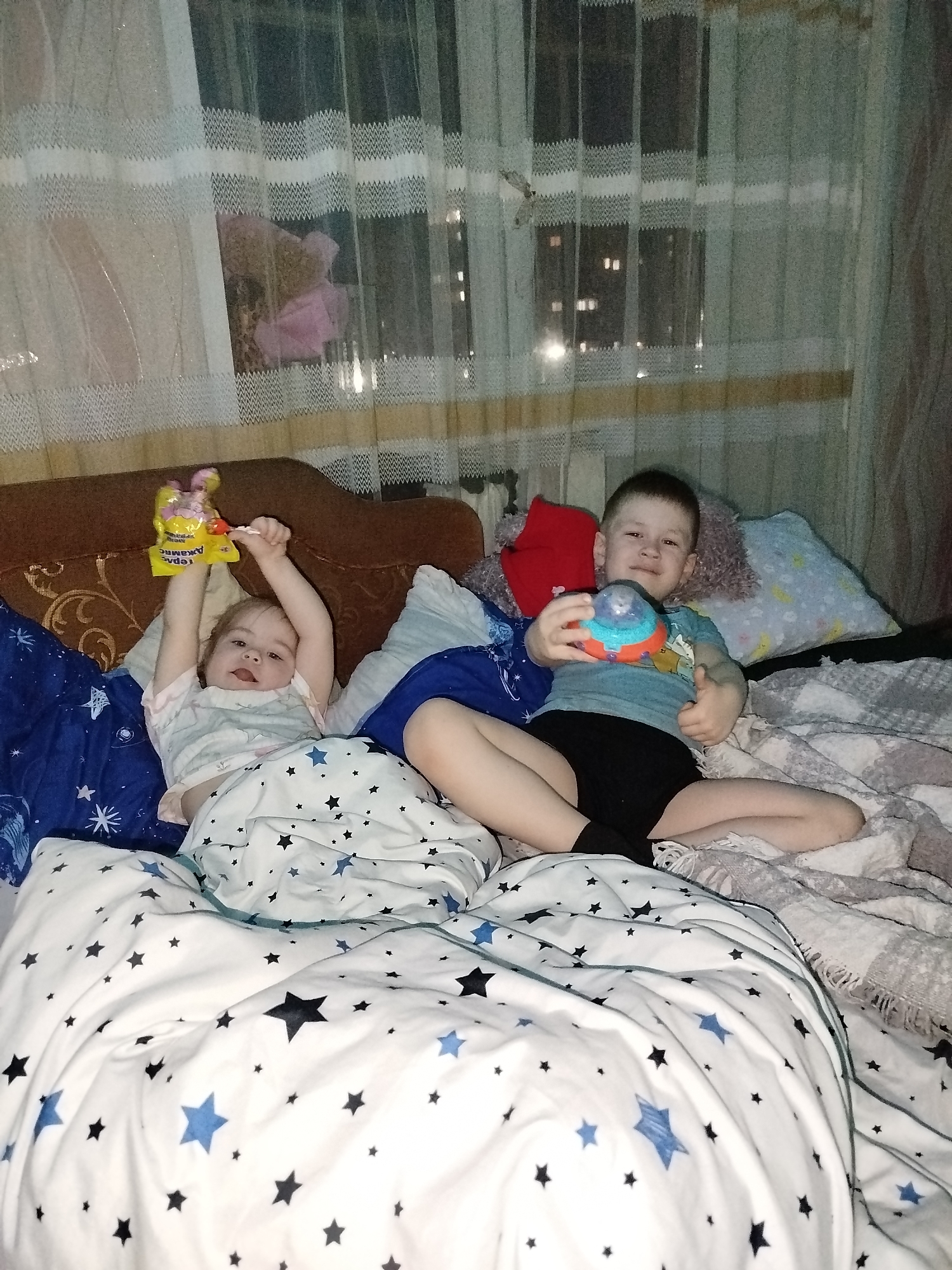 Location: Appears to be an indoor setting in an apartment, likely in Chaikovsky, Russia.
Scene: Two young children are lying on a bed. A younger child is on the left, holding a candy package with a lollipop, with one arm raised. The child is partially covered by a white duvet with a pattern of blue and black stars. The older child is on the right, sitting with crossed legs and holding a colorful toy. The bed has multiple pillows of varying colors and patterns. A brown headboard is visible behind the pillows. A window with sheer curtains and a view of buildings at night is in the background. The room lighting is dim.