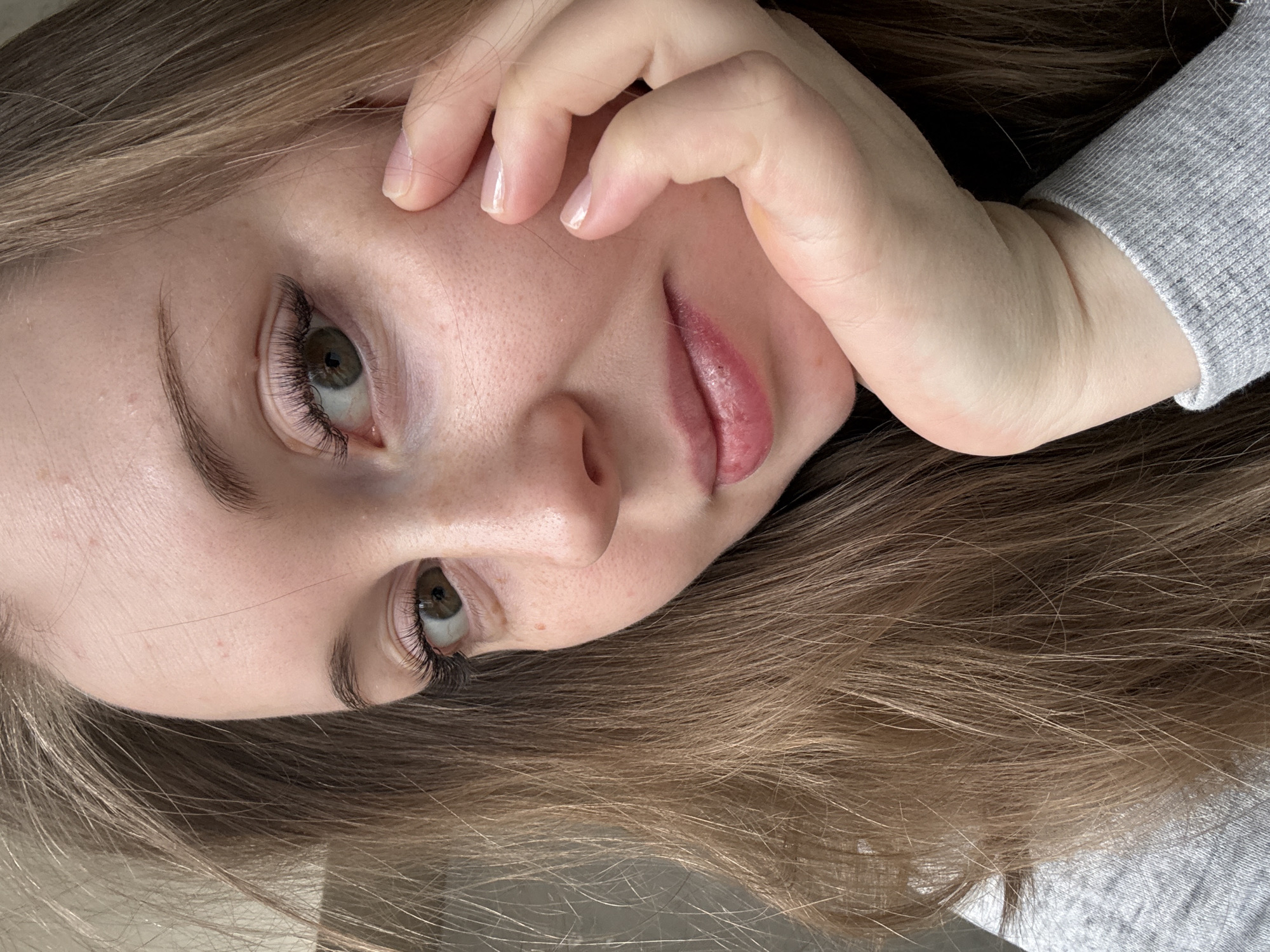 A close-up, angled view features a young woman's face, framed by light brown hair. She has fair skin, green eyes accentuated with dark eyelashes, and pink lips. Her right hand partially covers her face, with manicured fingernails visible. The woman is wearing a grey long-sleeved shirt or sweater. The background is slightly blurred. Location: Unknown, Unknown.